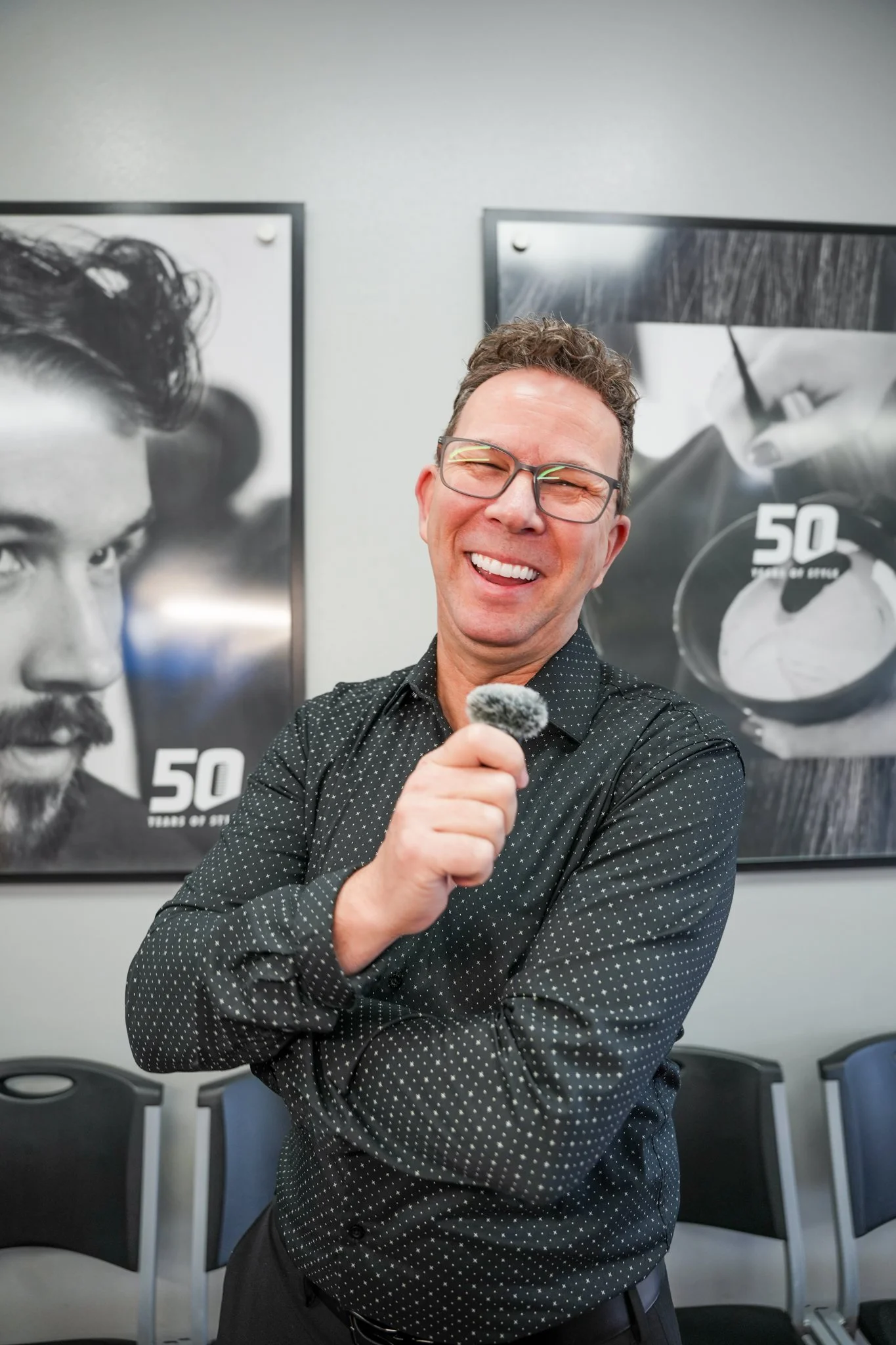 A man with glasses and a black shirt with white polka dots, smiling and holding a microphone, standing in front of black-and-white photographs on a wall