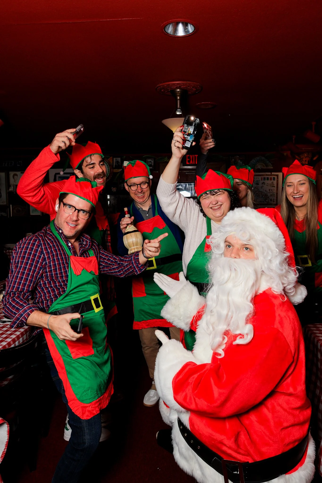 Group of people wearing Christmas-themed costumes and hats, taking a photo with Santa Claus in a festive setting.