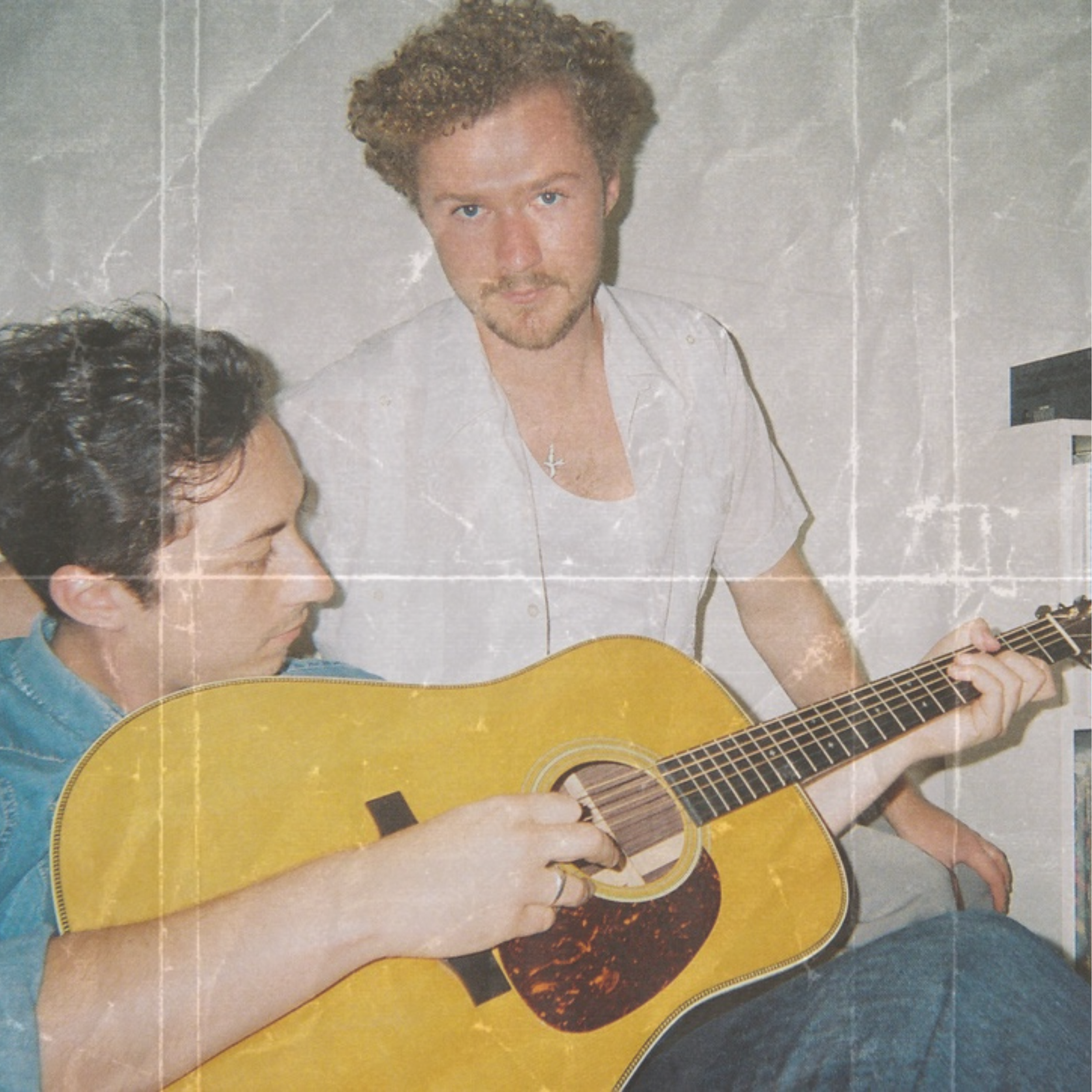 Two young men, one with curly hair playing an acoustic guitar, and the other with short dark hair watching, in a room with light-colored walls.