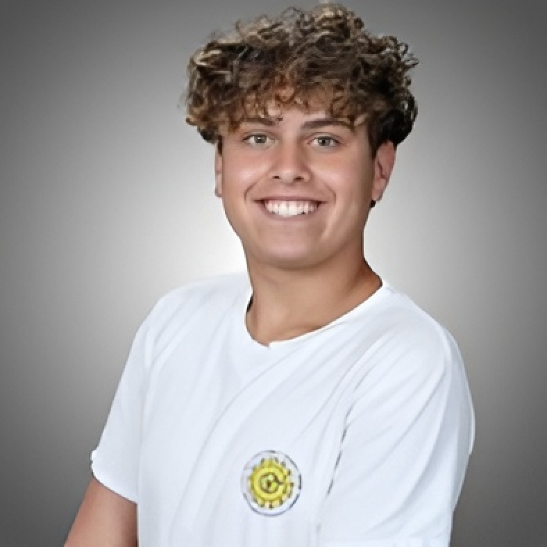 Young man with curly hair smiling, wearing a white T-shirt with a small yellow emblem on it, against a gray background.