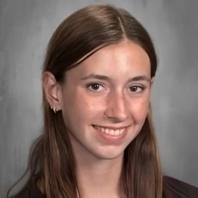 A young woman with light skin and long brown hair smiling at the camera against a plain gray background.