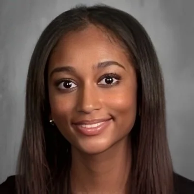 A young woman with long, straight brown hair and a warm smile, wearing a black top and earrings, against a gray background.