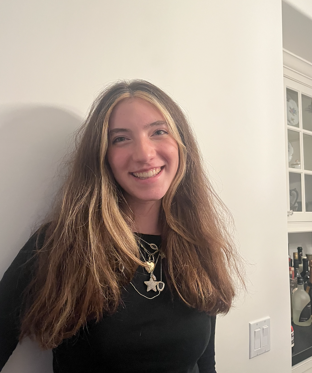 A young woman with long, wavy, light brown hair and wearing multiple necklaces, smiling at the camera. She is standing indoors near a white wall and a built-in cabinet.