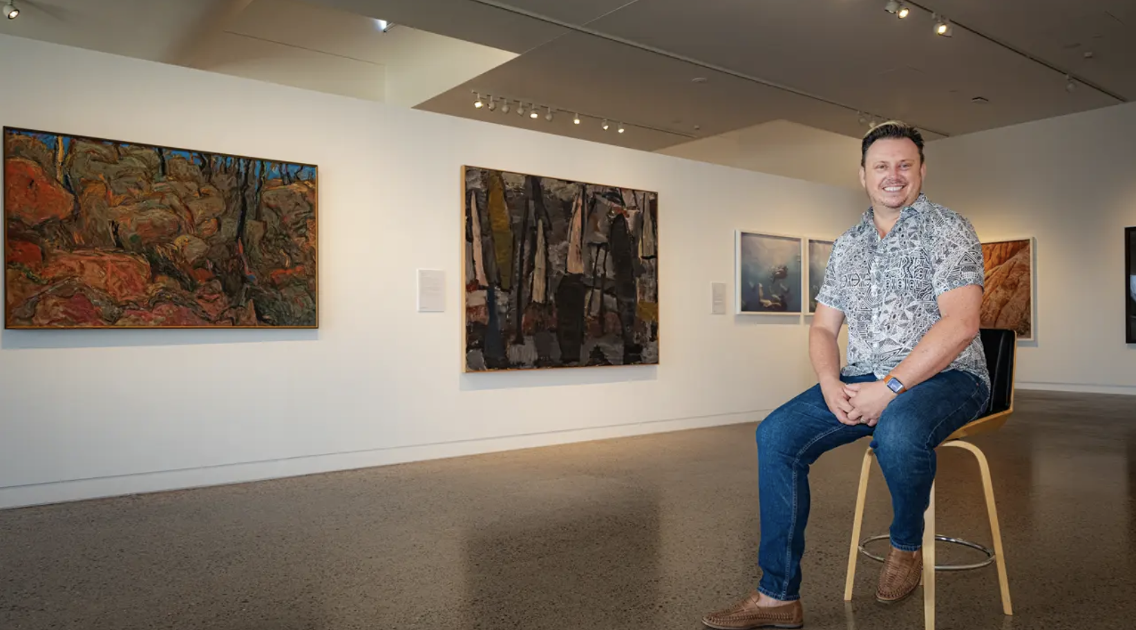 A man sitting on a chair in an art gallery, smiling, wearing a patterned short-sleeve shirt, jeans, and brown shoes, with several colorful paintings hanging on the white walls behind him.