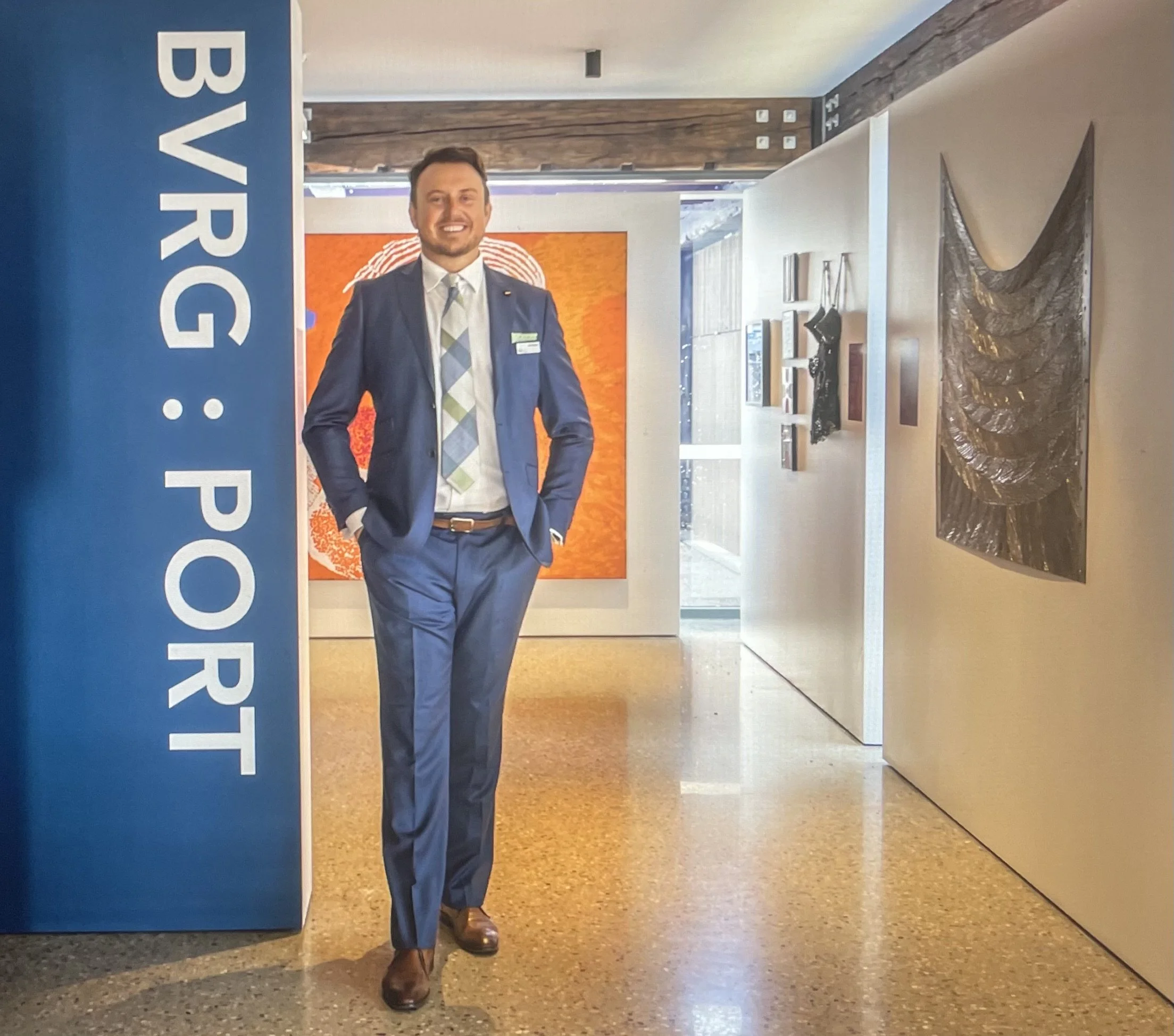 A man dressed in a blue suit and patterned tie standing next to a large sign that partially reads 'BUYER' in an indoor setting with art pieces and furniture.