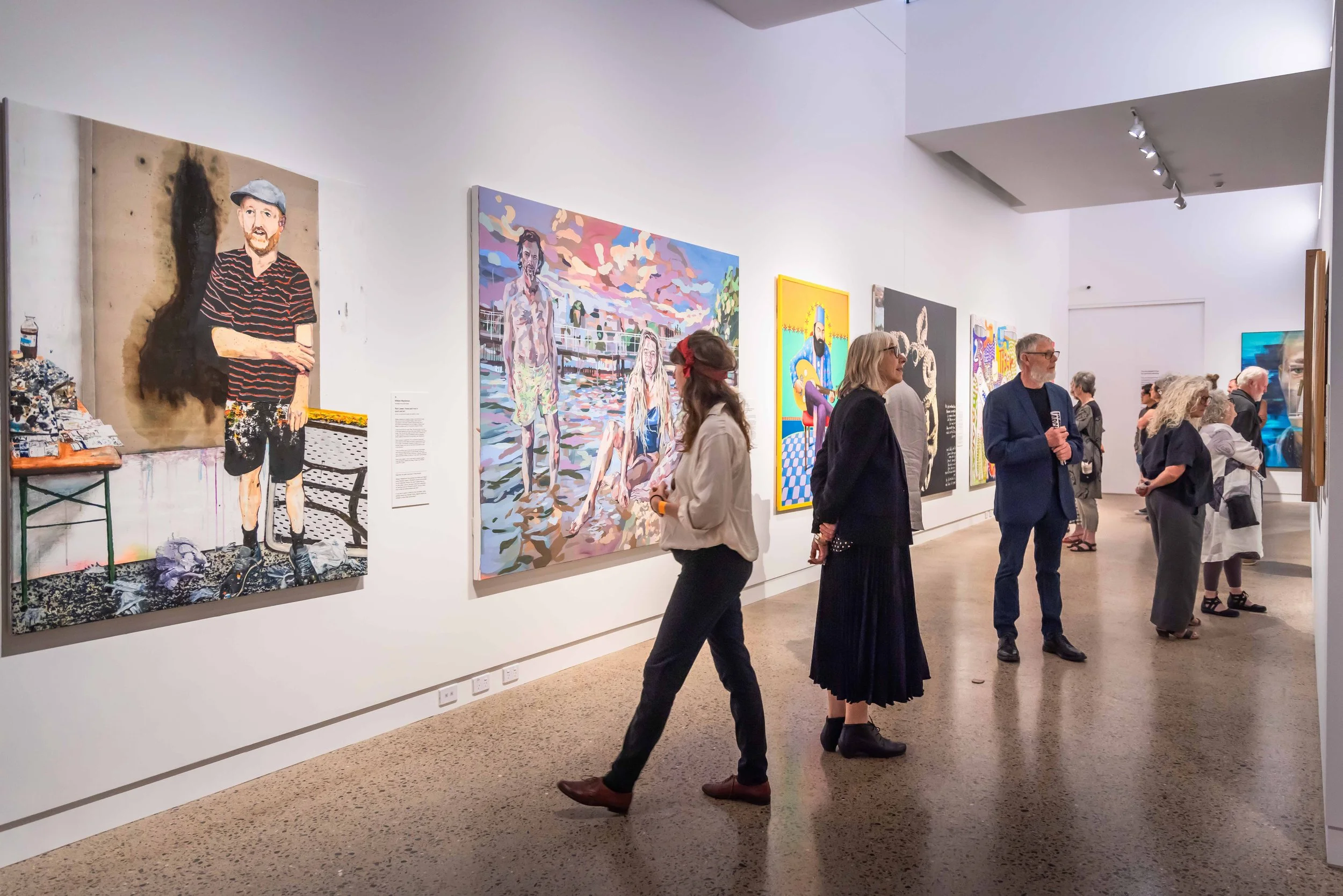 People viewing colorful artwork in an art gallery with white walls and track lighting. Various paintings, including a portrait of a bearded man and a family on a beach, are displayed on the wall.