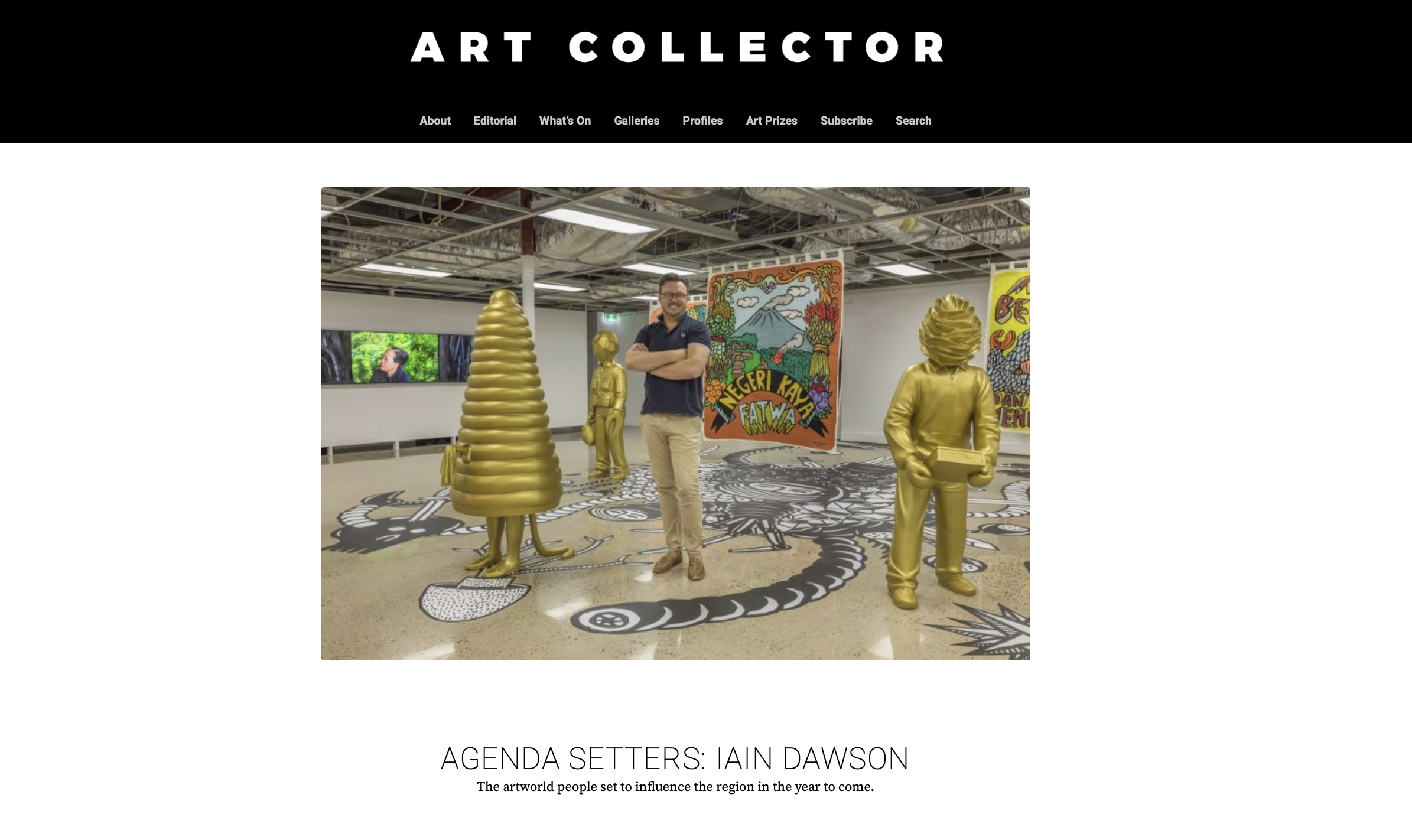 An art gallery display featuring four golden sculptures of human figures and objects, with colorful artwork and a TV screen in the background. A man stands in the center, smiling with arms crossed.