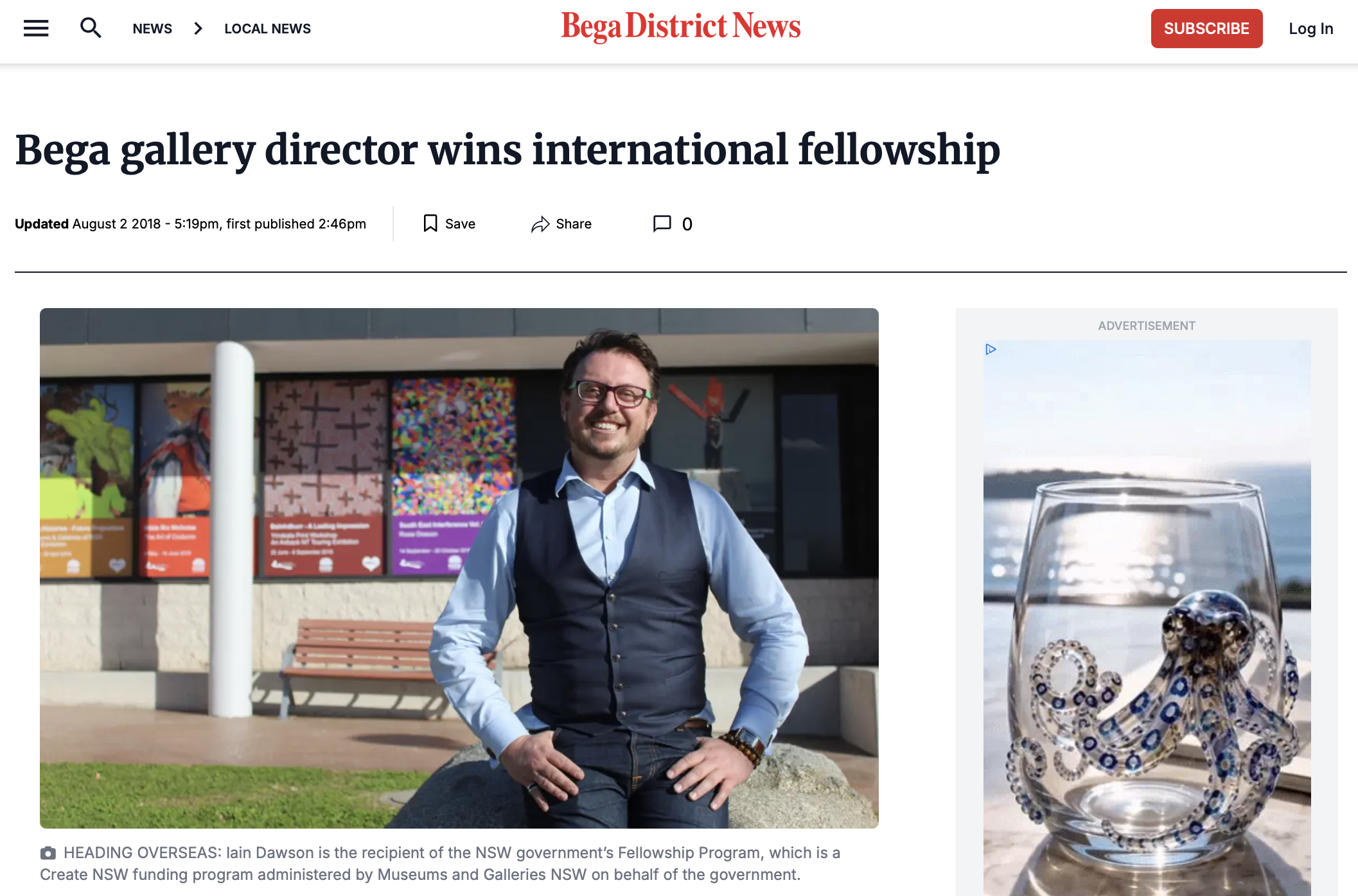 A man wearing glasses, a blue shirt, and a dark vest sitting on a rock outside a building with colorful posters displays at least part of a magazine or newspaper article about a gallery director named Iain Dawson. In the background, a building features artwork posters, and to the right, a glass with jewelry inside is partially visible.
