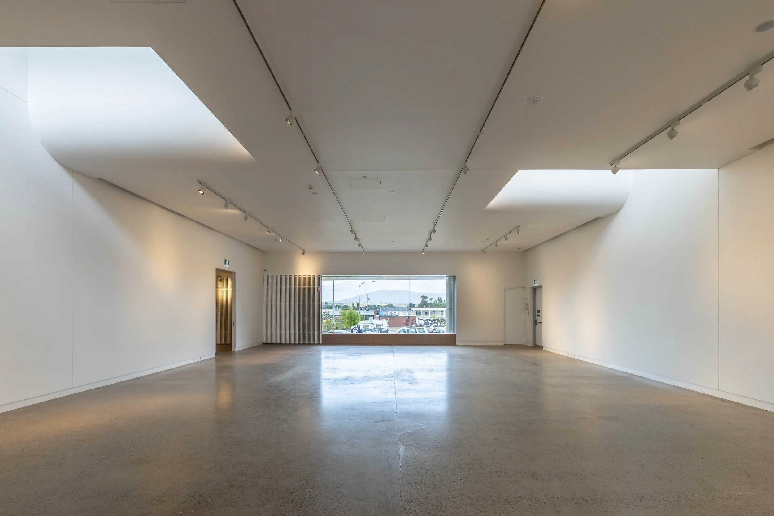 Interior view of a spacious, minimalistic room with white walls, a large window at the back letting in natural light, and track lighting on the ceiling.