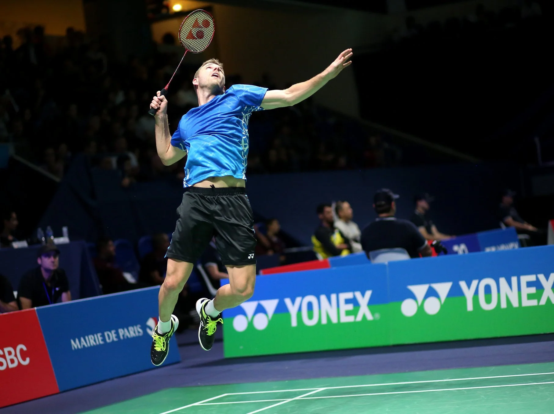 Badminton player in blue shirt jumping and preparing to hit a shuttlecock with a racket on a Yonex-branded court.