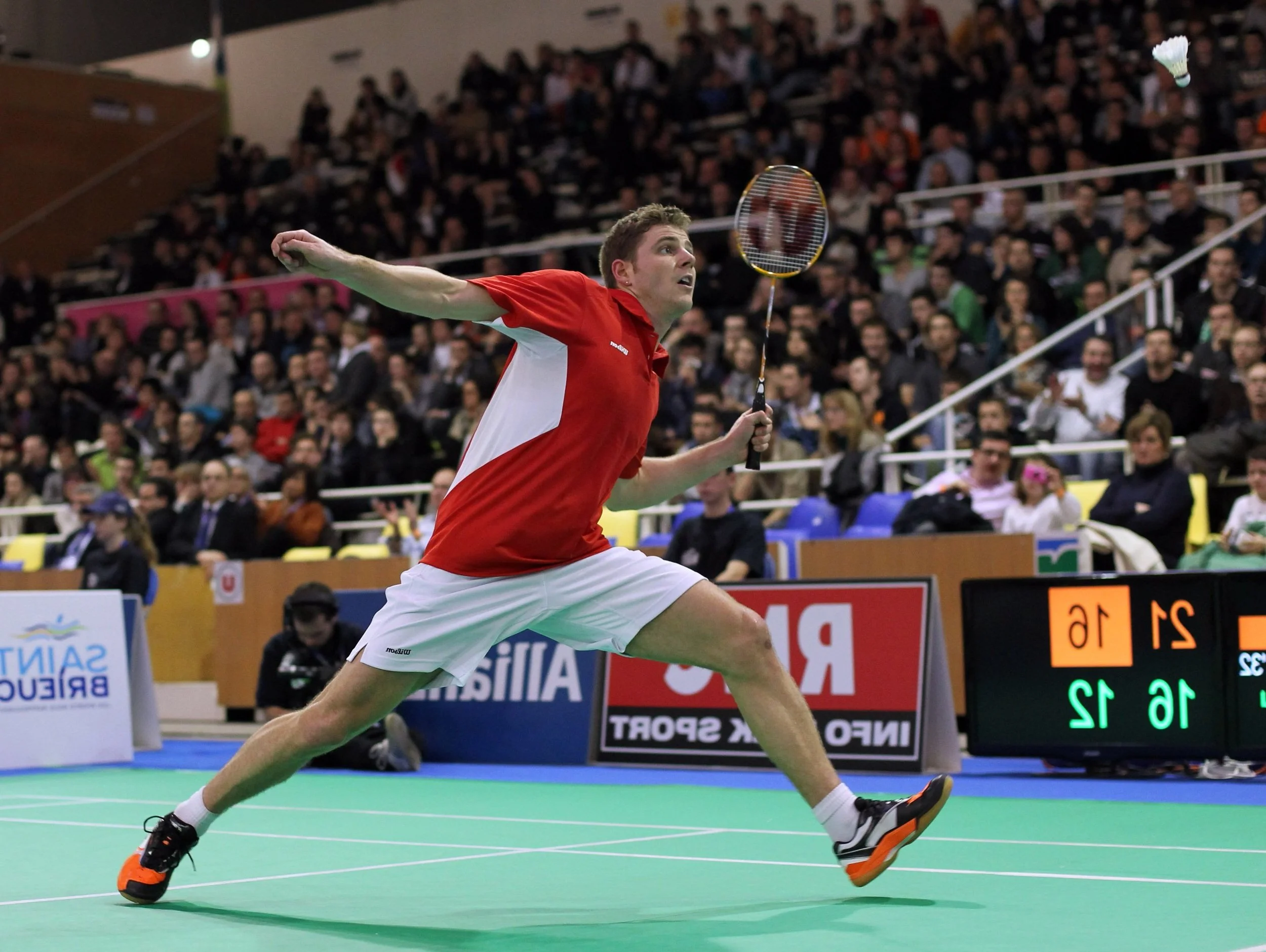 A male badminton player in a red and white outfit lunges towards a shuttlecock on a court during a match, with an audience watching in the background.