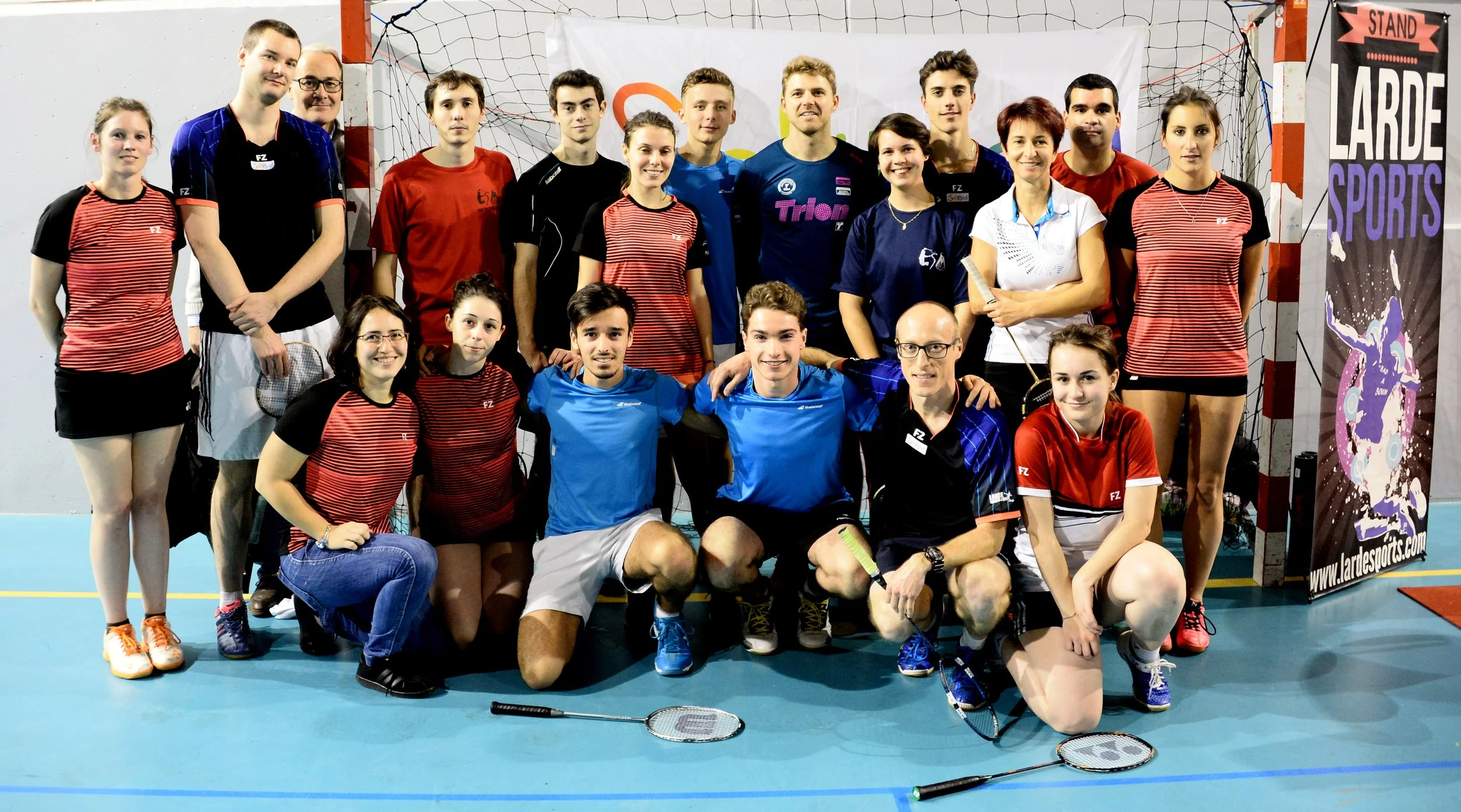 Group photo of badminton players posing in a sports hall, holding rackets with a "Larde Sports" banner in the background.