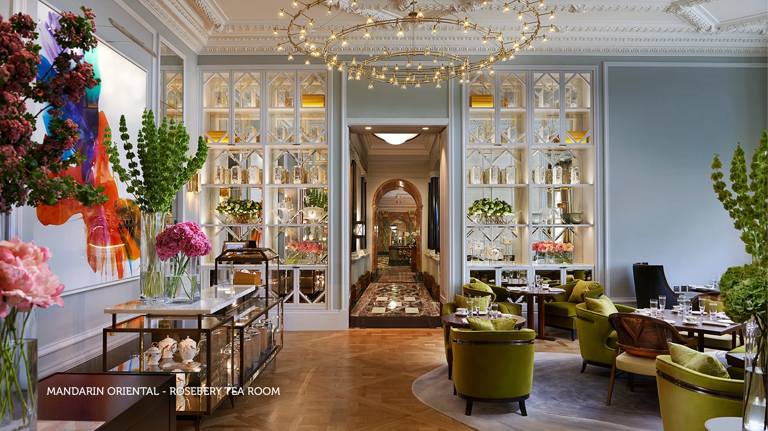 Elegant hotel lounge with green velvet armchairs around a circular rug, large vases with flowers, glass display shelves, and ornate ceiling with chandelier lighting.