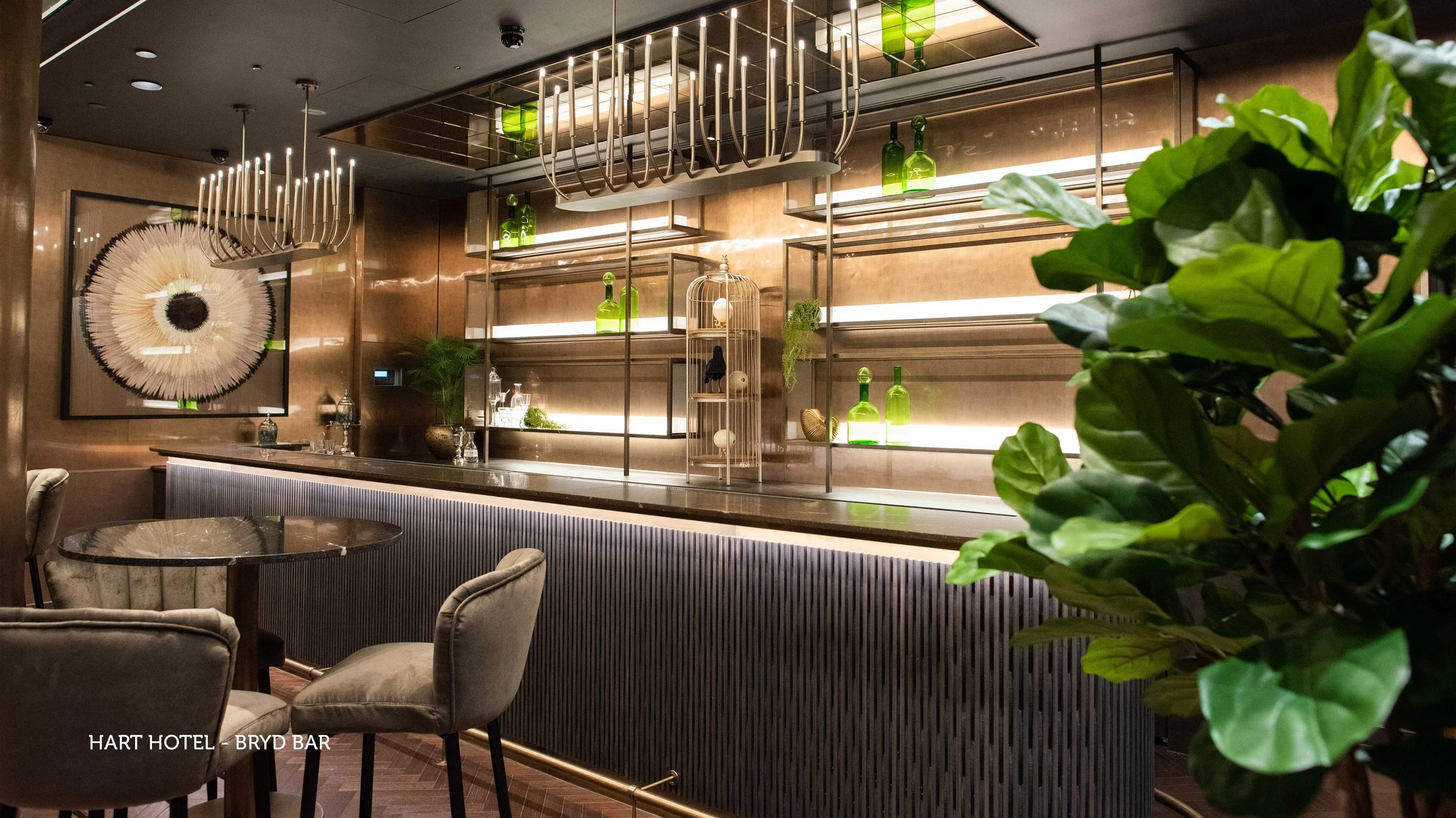 Elegant bar with modern decor, dark wood flooring, a black ribbed bar counter, green velvet chairs around a small round table, and a well-stocked back bar with bottles and glassware. Decor includes a large wall art piece and green plants.