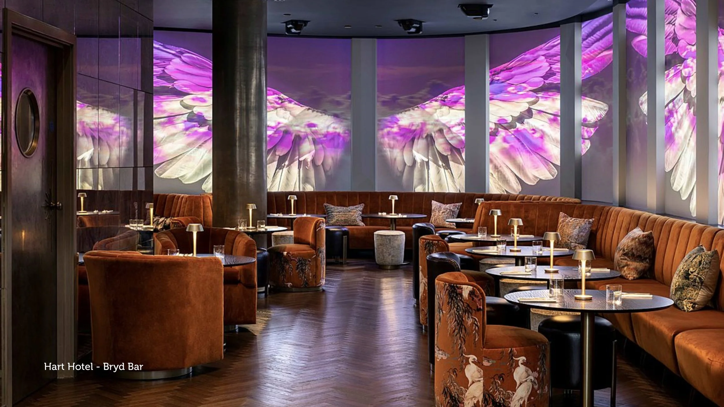 A stylish lounge with curved seating, small tables with lamps, patterned chairs, and a digital wall display of pink and purple flowers.