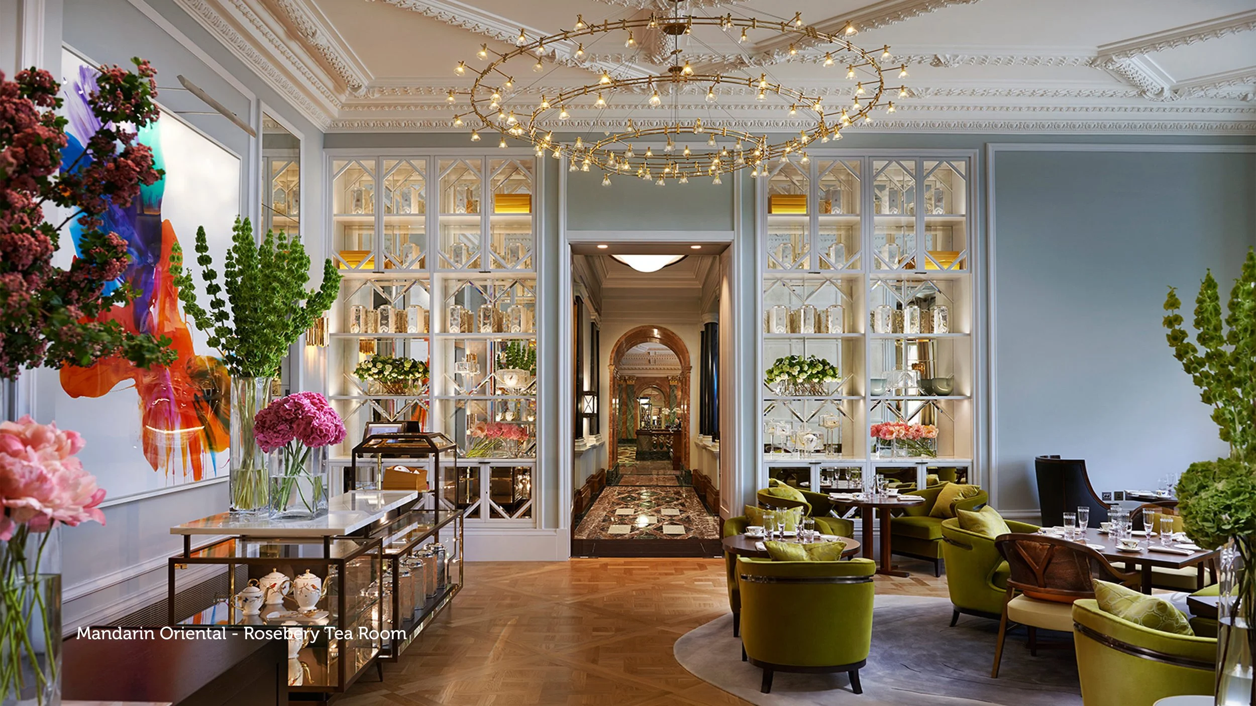 Elegant hotel lounge with green velvet armchairs around a circular rug, large vases with flowers, glass display shelves, and ornate ceiling with chandelier lighting.