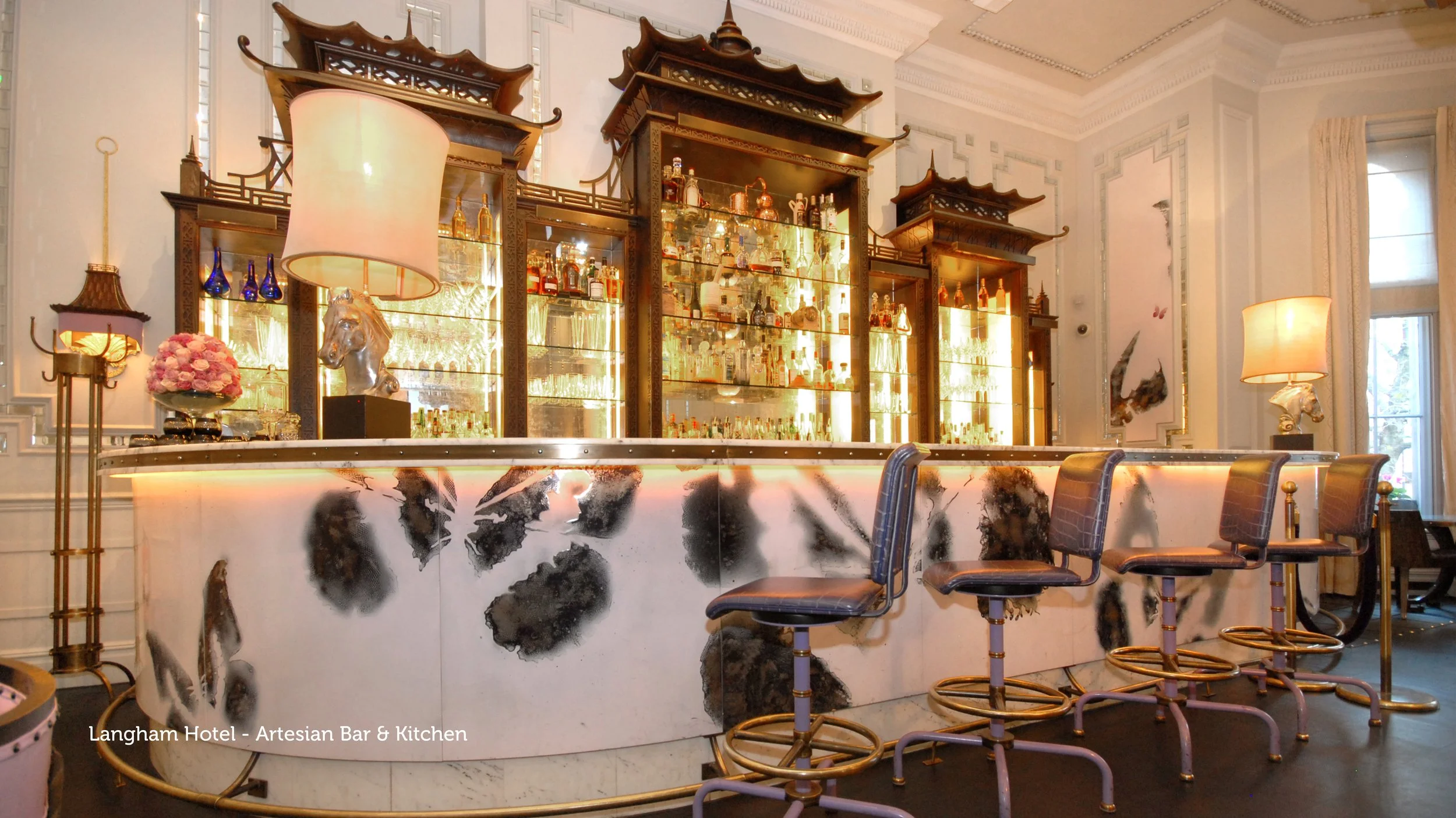 Elegant bar with a marble front, illuminated shelves filled with bottles, decorative lamps, and leather bar stools in a luxurious setting.