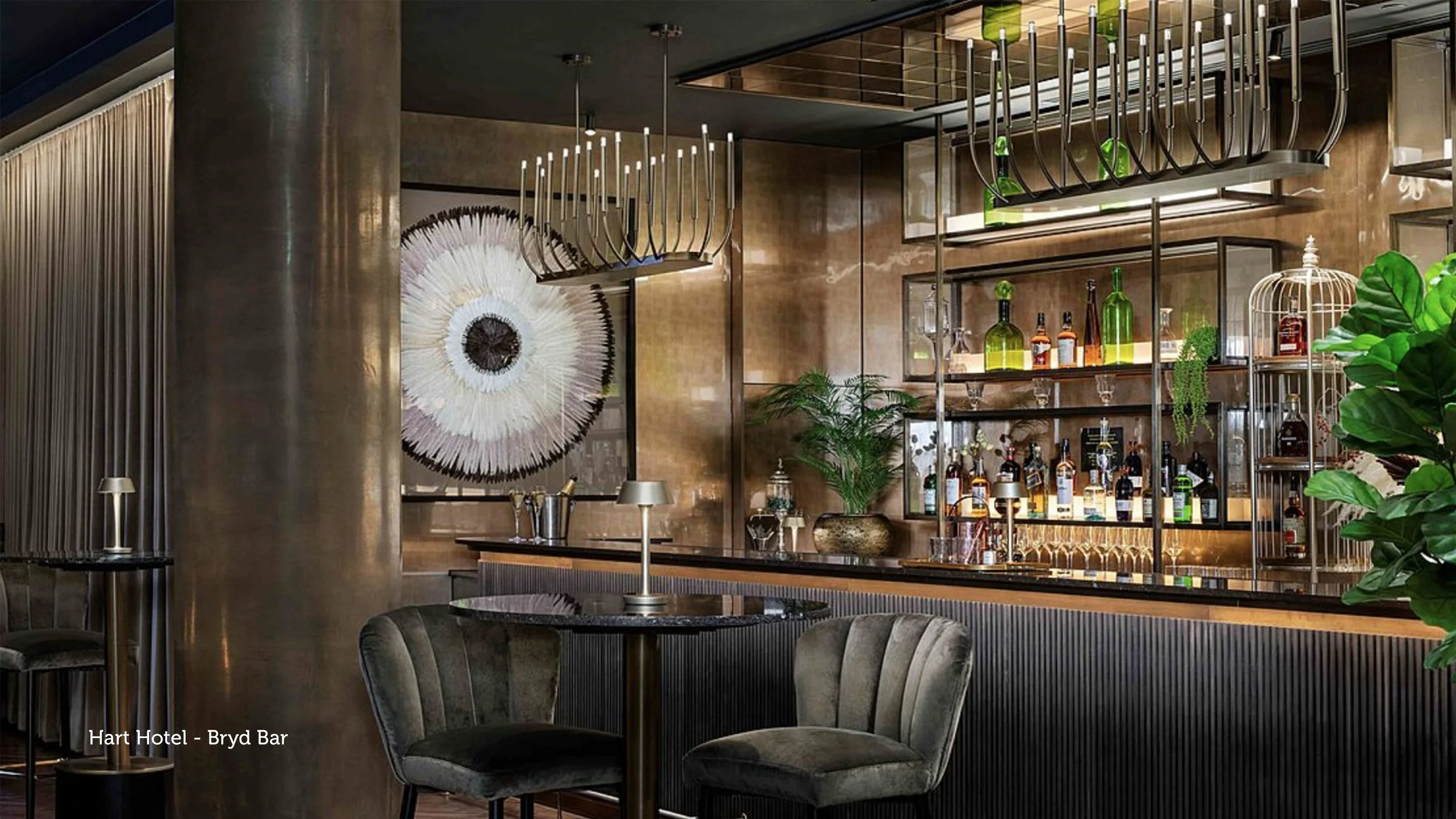 Elegant bar with modern decor, dark wood flooring, a black ribbed bar counter, green velvet chairs around a small round table, and a well-stocked back bar with bottles and glassware. Decor includes a large wall art piece and green plants.