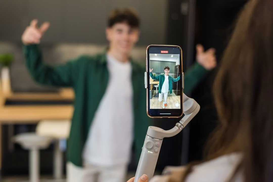 Junge Person beim Aufnehmen eines Videos mit Smartphone und Gimbal stabilisiert, im Hintergrund Studenten in einem modernen Raum