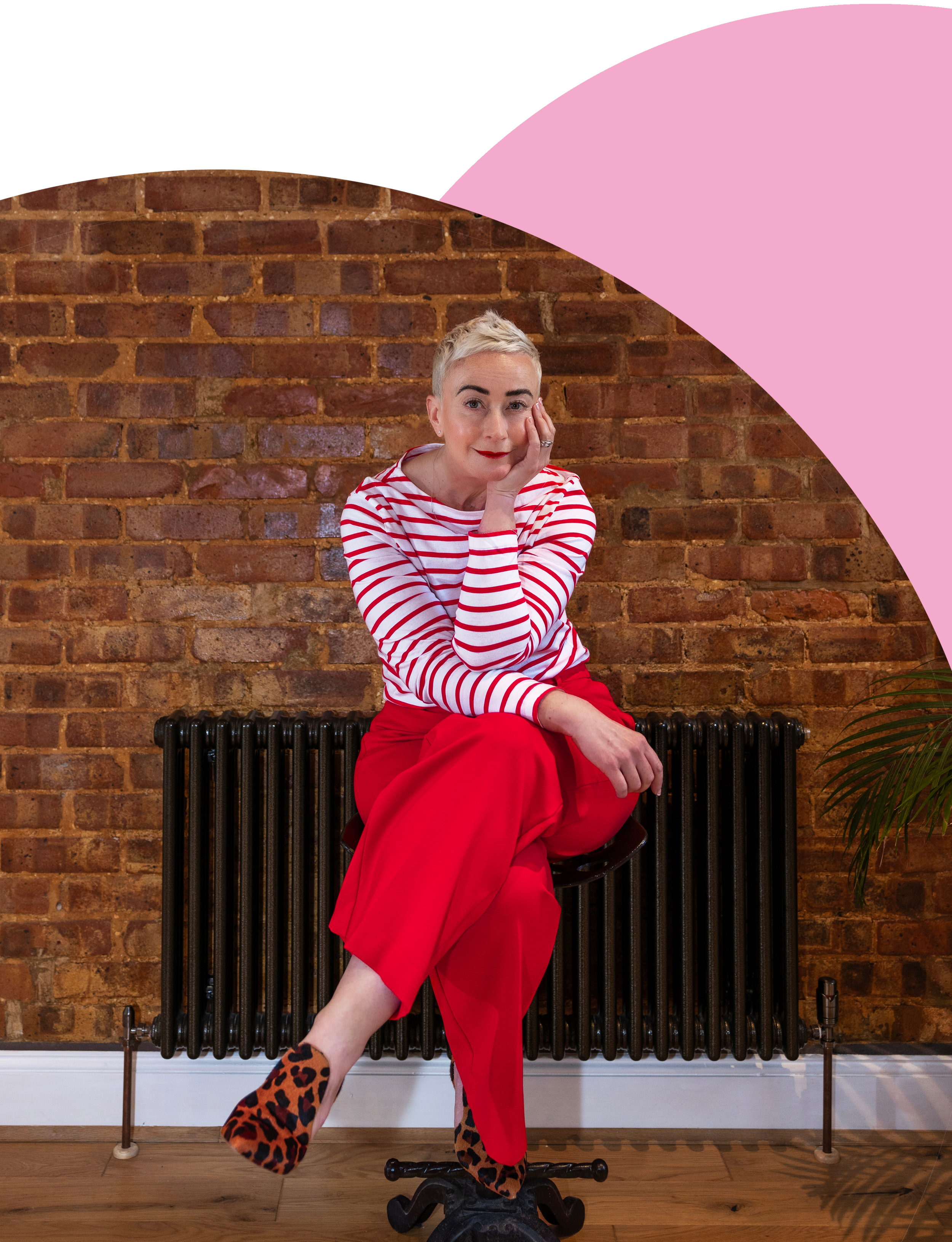 A woman with short blonde hair, wearing a red and white striped long-sleeve shirt, red pants, and leopard print shoes, sitting on a black radiator in front of a brick wall. She is resting her face on her hand, looking at the camera with a slight smile.