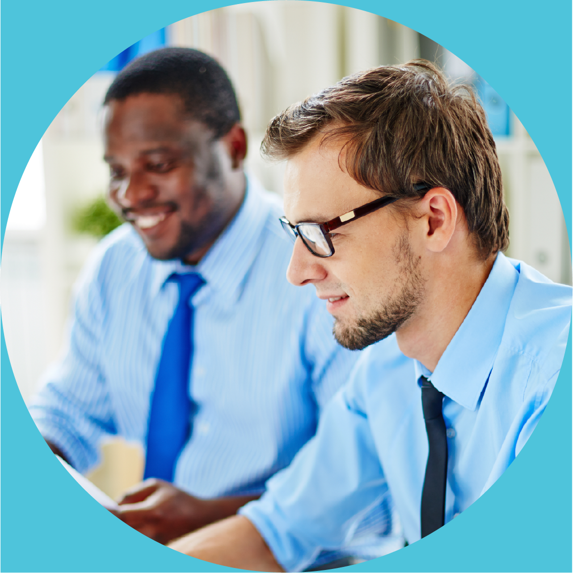 Two men in business attire sitting at a desk, smiling, in an office environment.