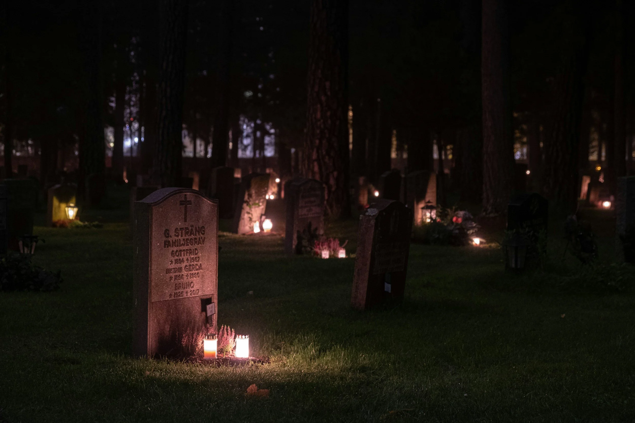 Nighttime scene of a cemetery with gravestones, illuminated by candles and small lights, surrounded by tall trees.