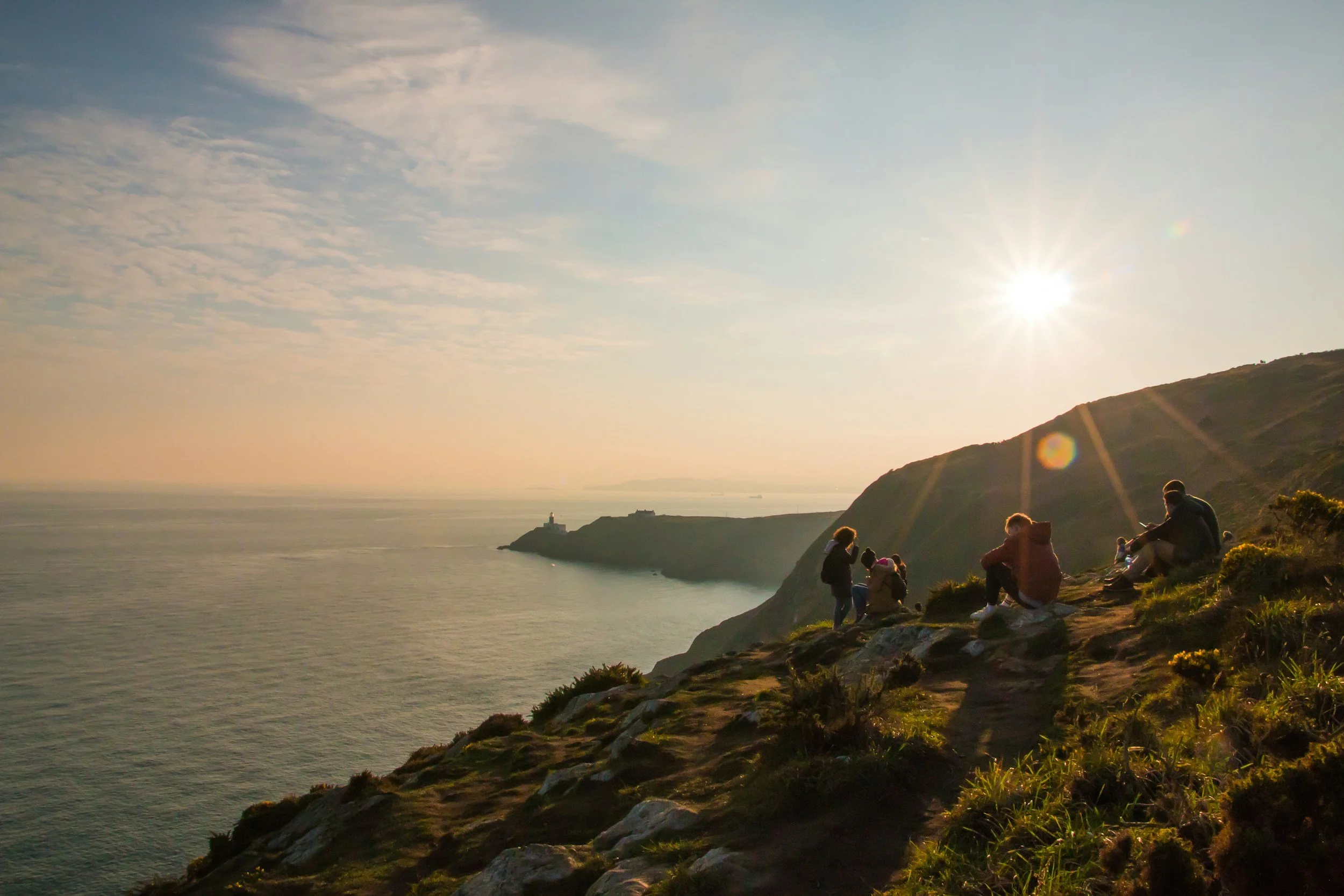 People sitting and standing on a grassy cliffside watching the sunset over the ocean with a lighthouse in the distance.