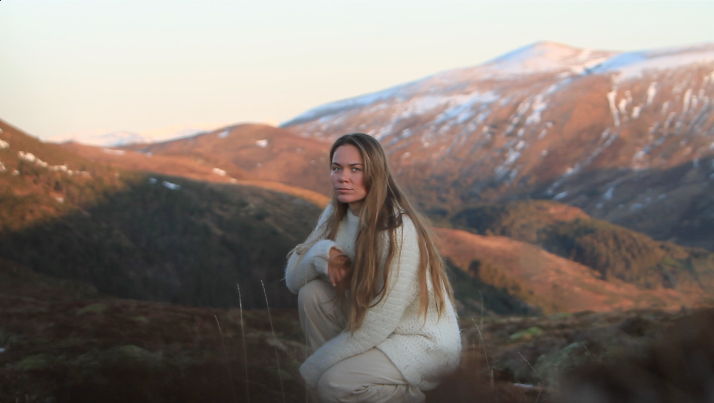 A woman with long hair in a cream sweater and light-colored pants crouches in a mountainous landscape with snow-capped peaks in the background, gazing at the camera.