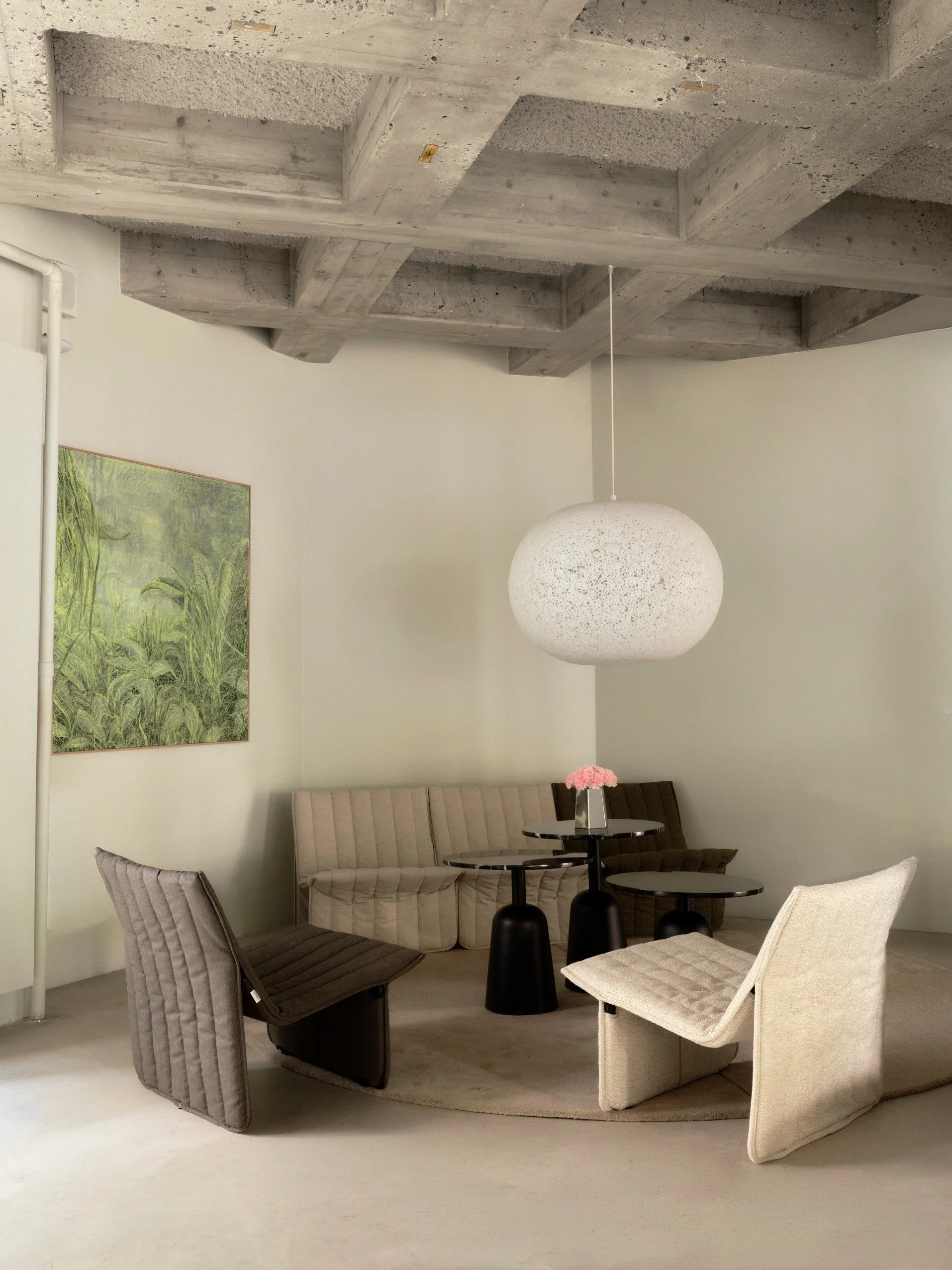 A living room with modern furniture, including a beige sofa and colorful chairs surrounding black and beige side tables. A large, spherical white pendant light hangs from the ceiling. There is a framed botanical print on the wall and a vase with pink flowers on one of the tables.