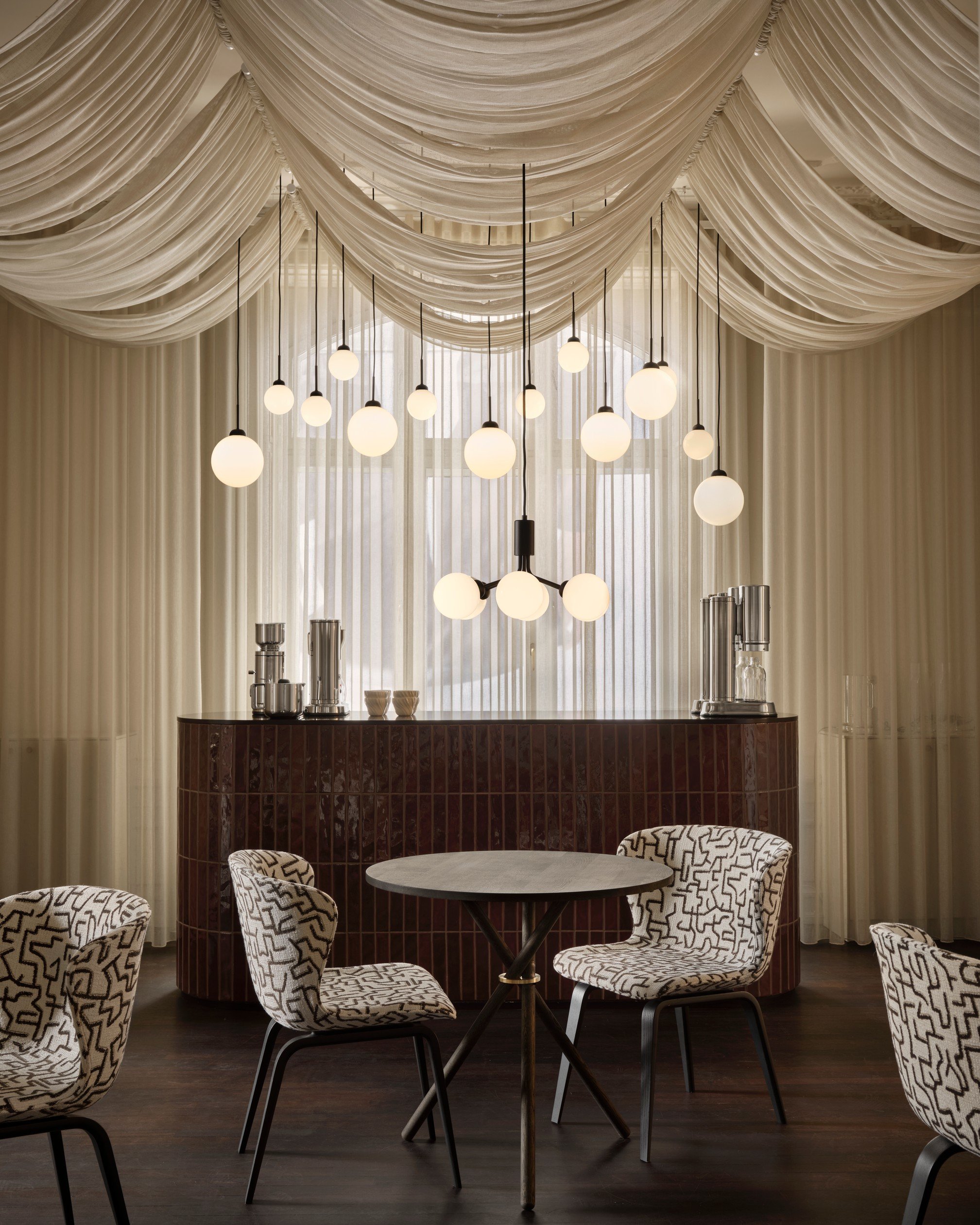 Elegant dining area with a marble round table surrounded by patterned upholstered chairs, a velvet curtain, and a marble bar with modern pendant lights overhead.