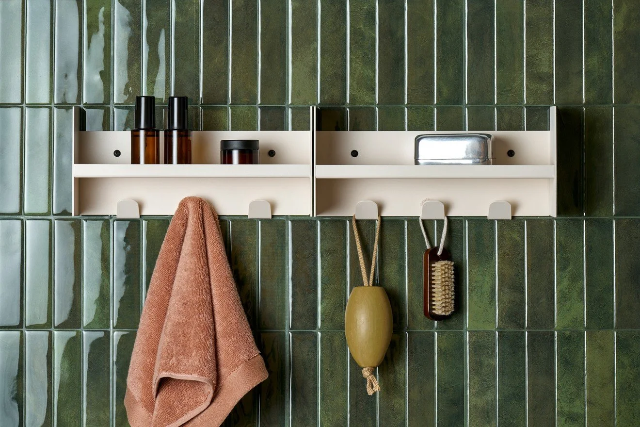 A white wall-mounted shelf with toiletries above hooks on a green tiled bathroom wall. The hooks hold a pink towel, a yellow bathbrush, and a small scrub brush.