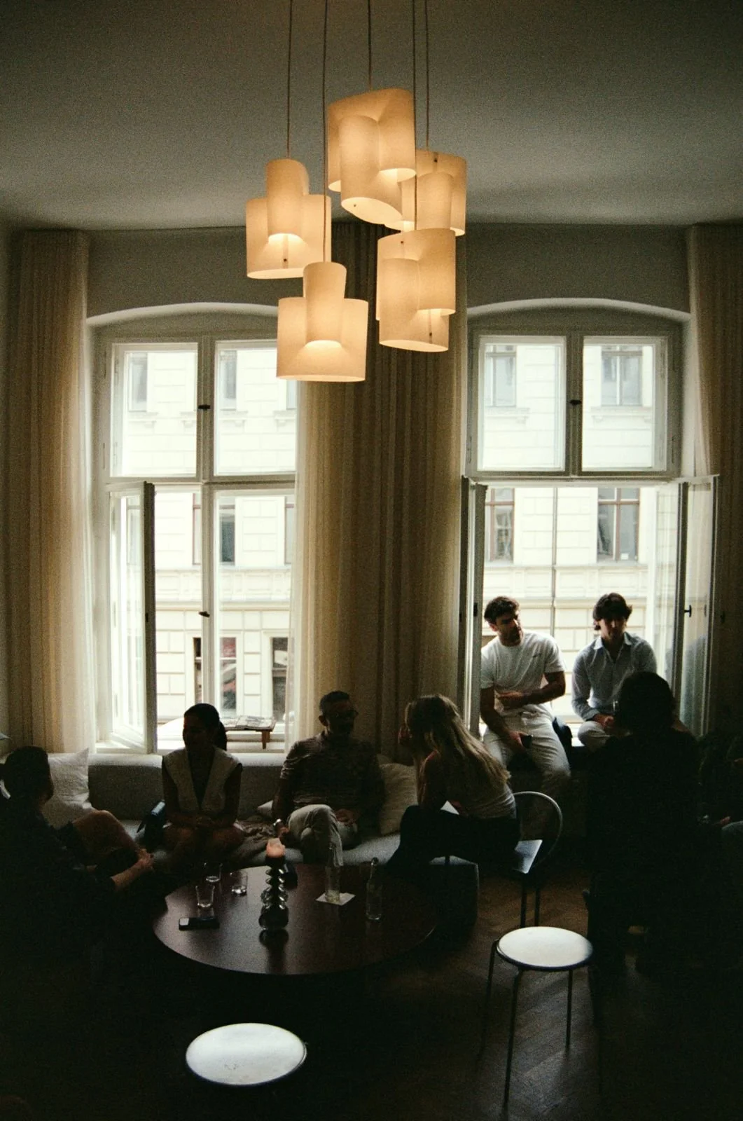 Group of people socializing in a dimly lit living room with large windows, a modern chandelier, and a sitting area.