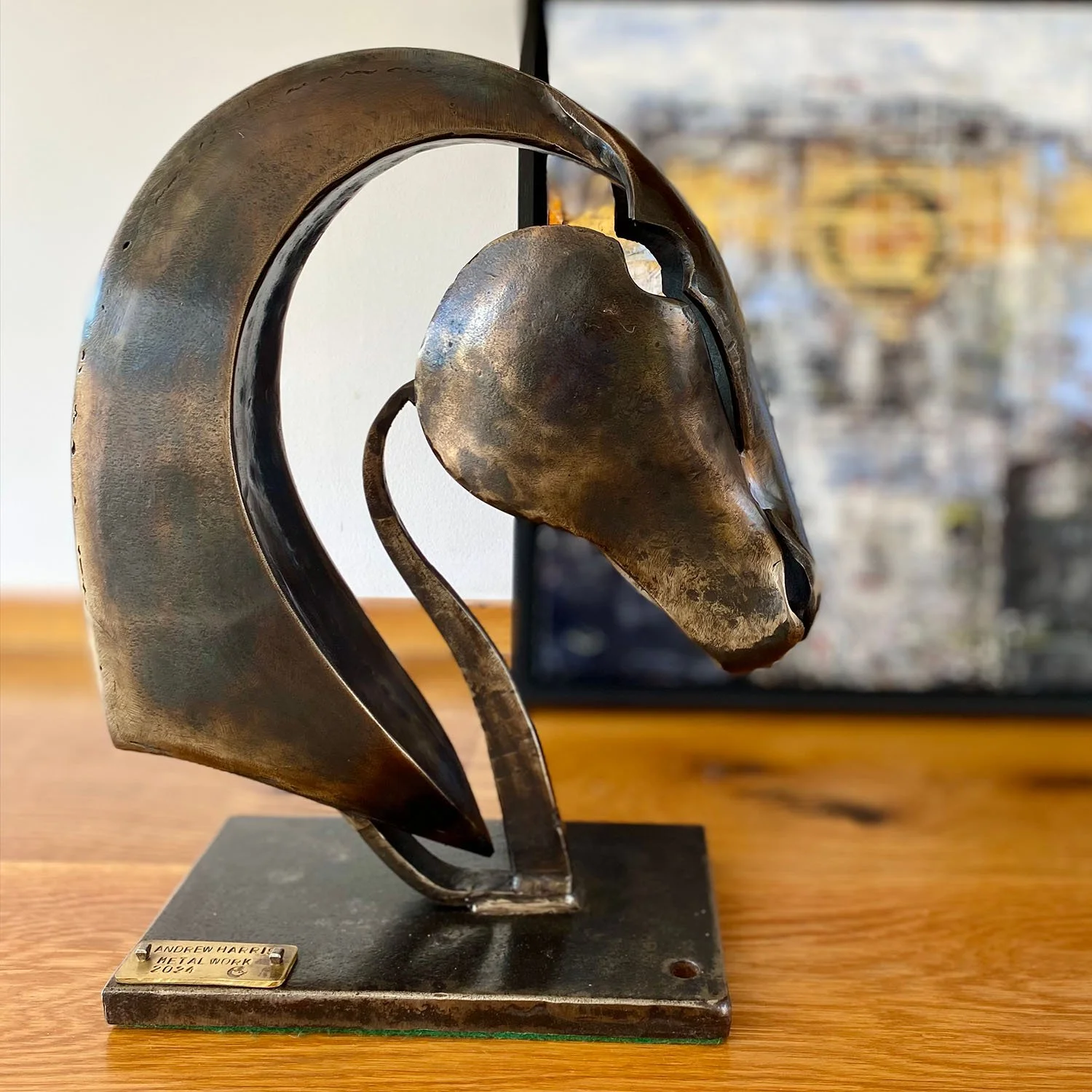 A metal forged sculpture of horses head in art gallery with blurred abstract painting in the background.