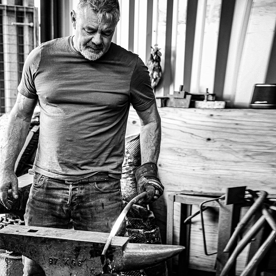 Black and white image of man in metalworkers workshop working a red hot piece of metal on anvil.