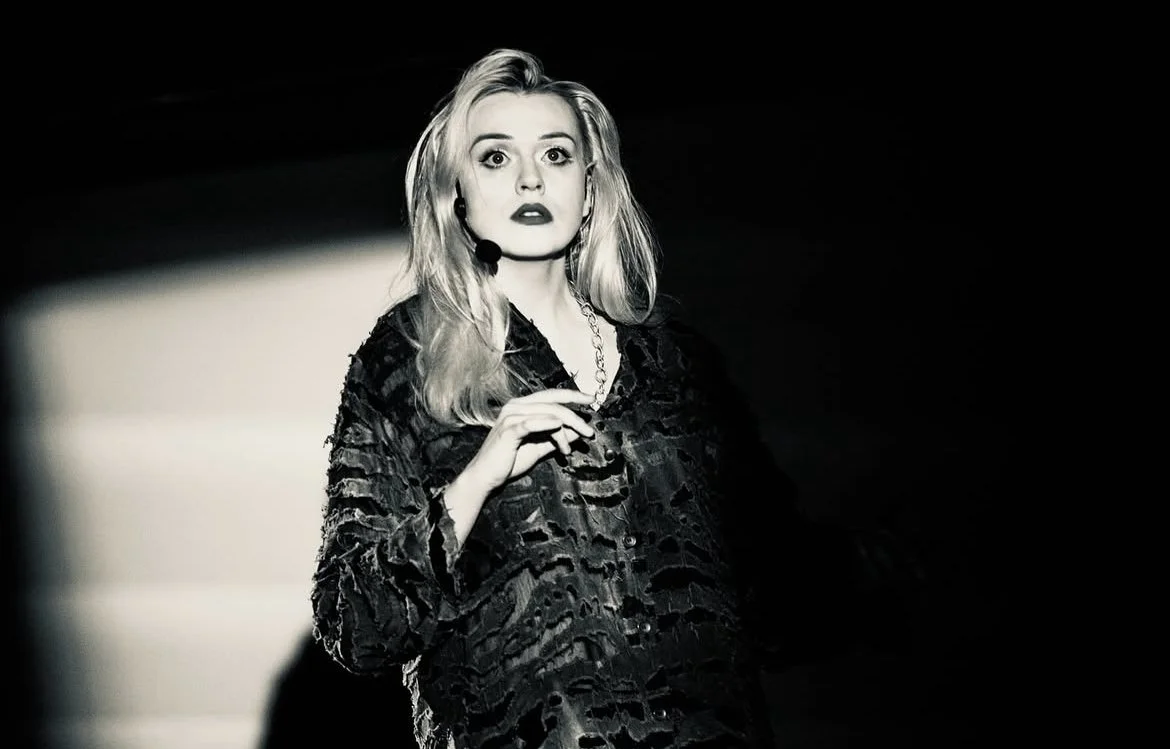 Salomeja Petronyte performing Bronius Kutavičius' "Mažasis spektkalis“ (the little performance) with the Fluorescence ensemble in Lithuania, the singer wears a black dress, black and white colour picture, blond hair.