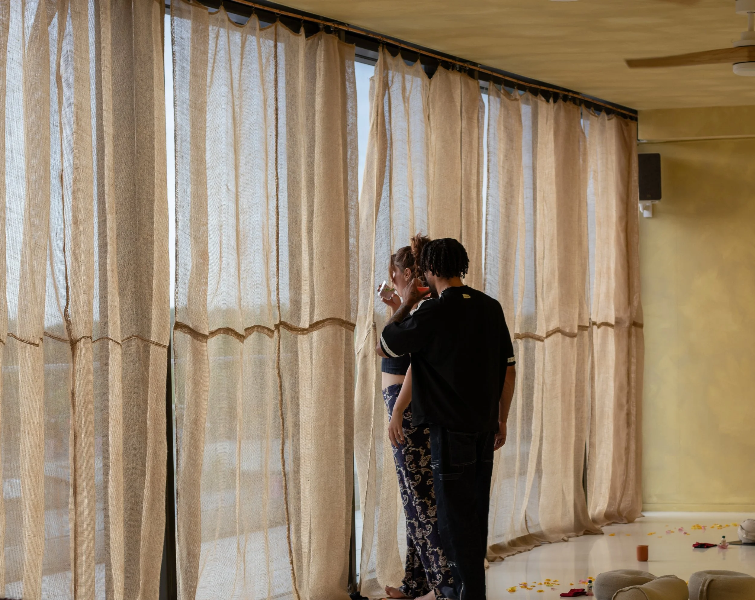 Two people standing in front of beige curtains, with one drinking from a glass while the other stands nearby.