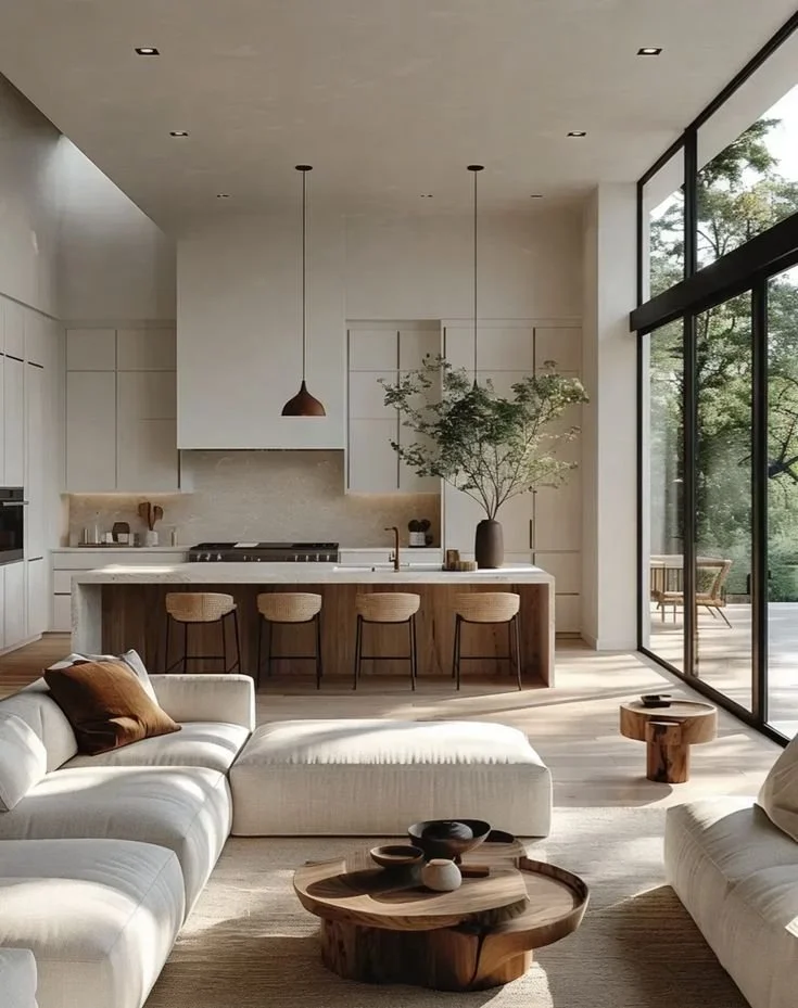 Modern open-concept living room and kitchen with large windows, white furniture, natural wood accents, and a green plant