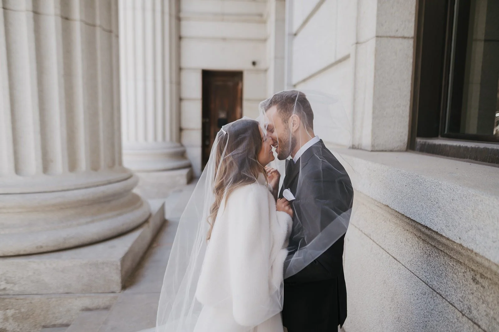 Bride and groom sharing a kiss under the veil against stone columns in winter