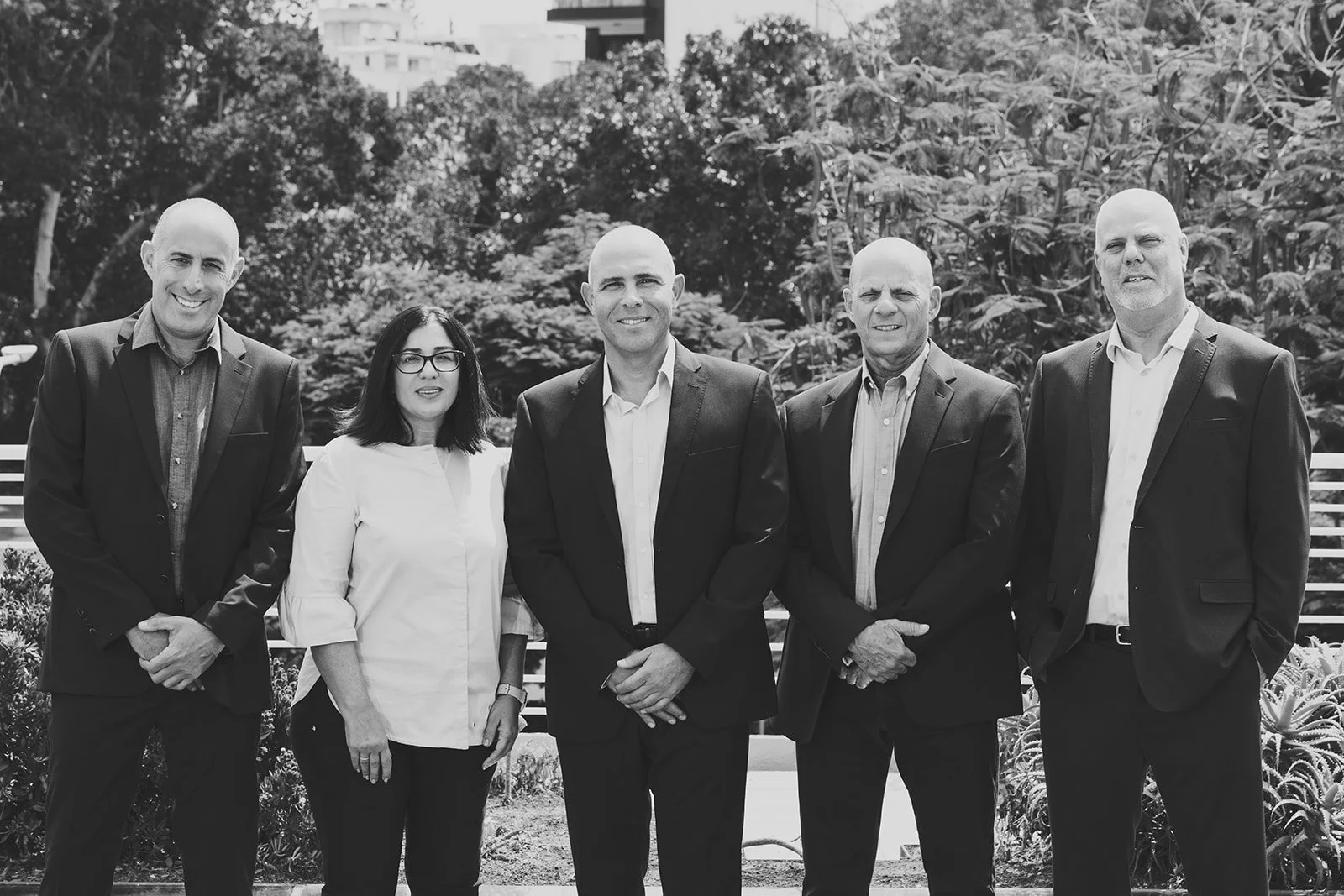 Group of five professionally dressed individuals, four men and one woman, standing outdoors with trees in the background. All are facing forward and smiling.
