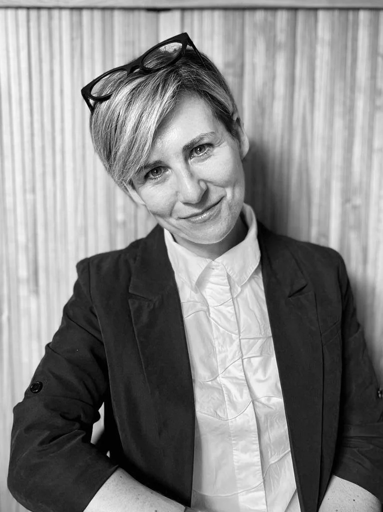 Black and white photo of a woman with short blonde hair, wearing glasses on top of her head, a white shirt, and a dark blazer, sitting in front of a wooden wall.