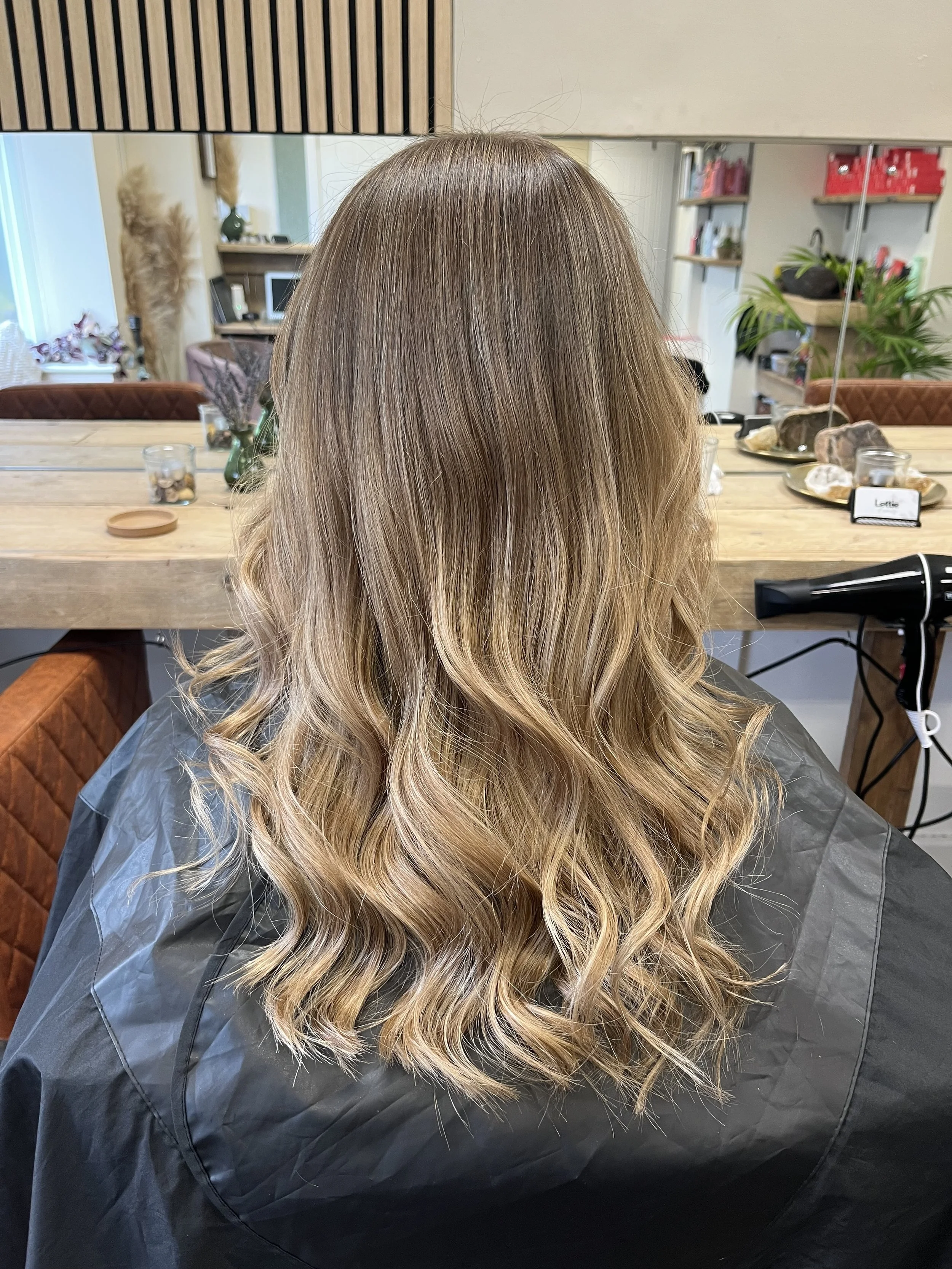 Back view of a woman with medium-length, wavy, blonde hair sitting in a salon chair.