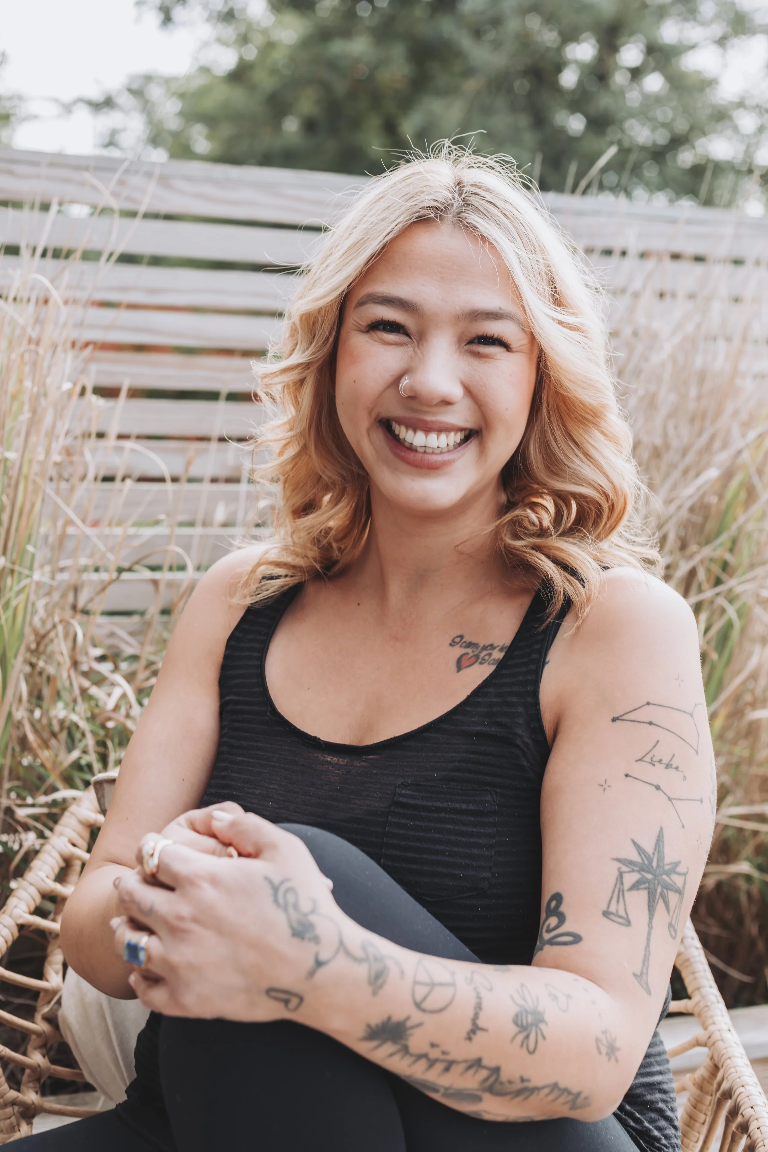 Smiling woman with wavy blonde hair, wearing a sleeveless black top, sitting outdoors with her arms crossed, tattoos on her arms and chest, and a background of bushes and a wooden fence.