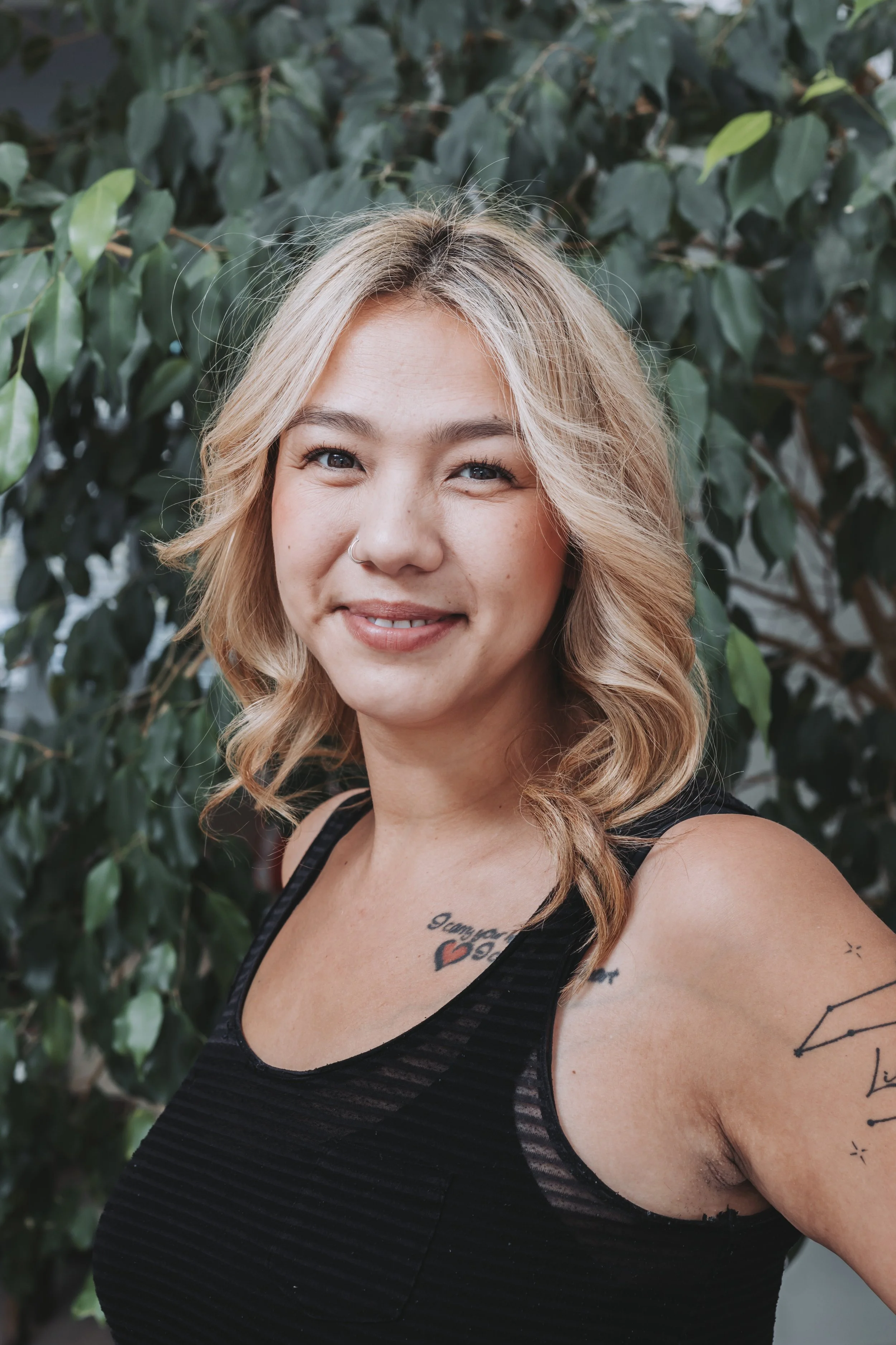 A woman with blonde wavy hair, a nose piercing, and tattoos on her shoulder and chest, smiling while standing in front of green leafy plants.