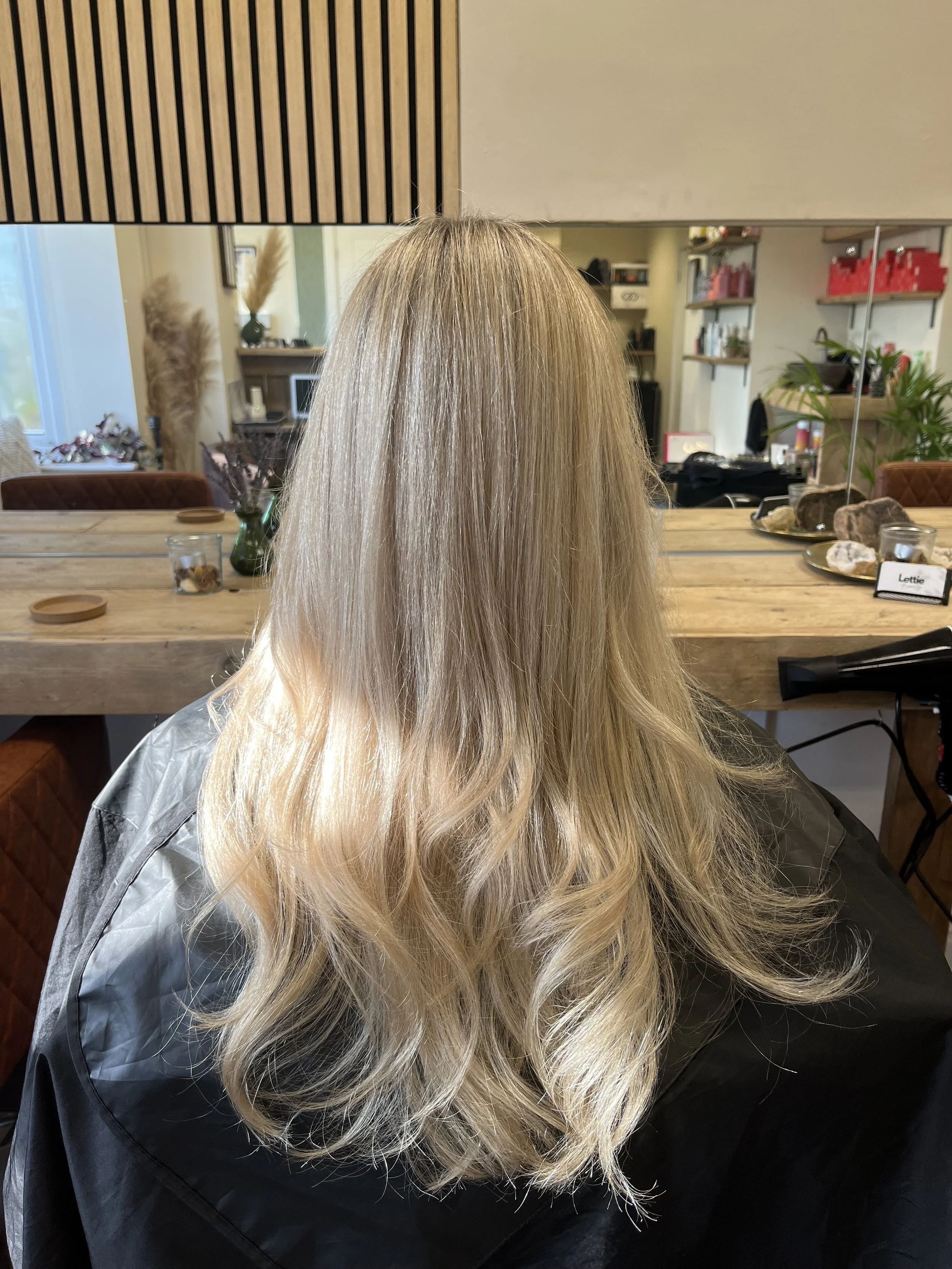 Back view of a woman with long blonde wavy hair seated in a hair salon.