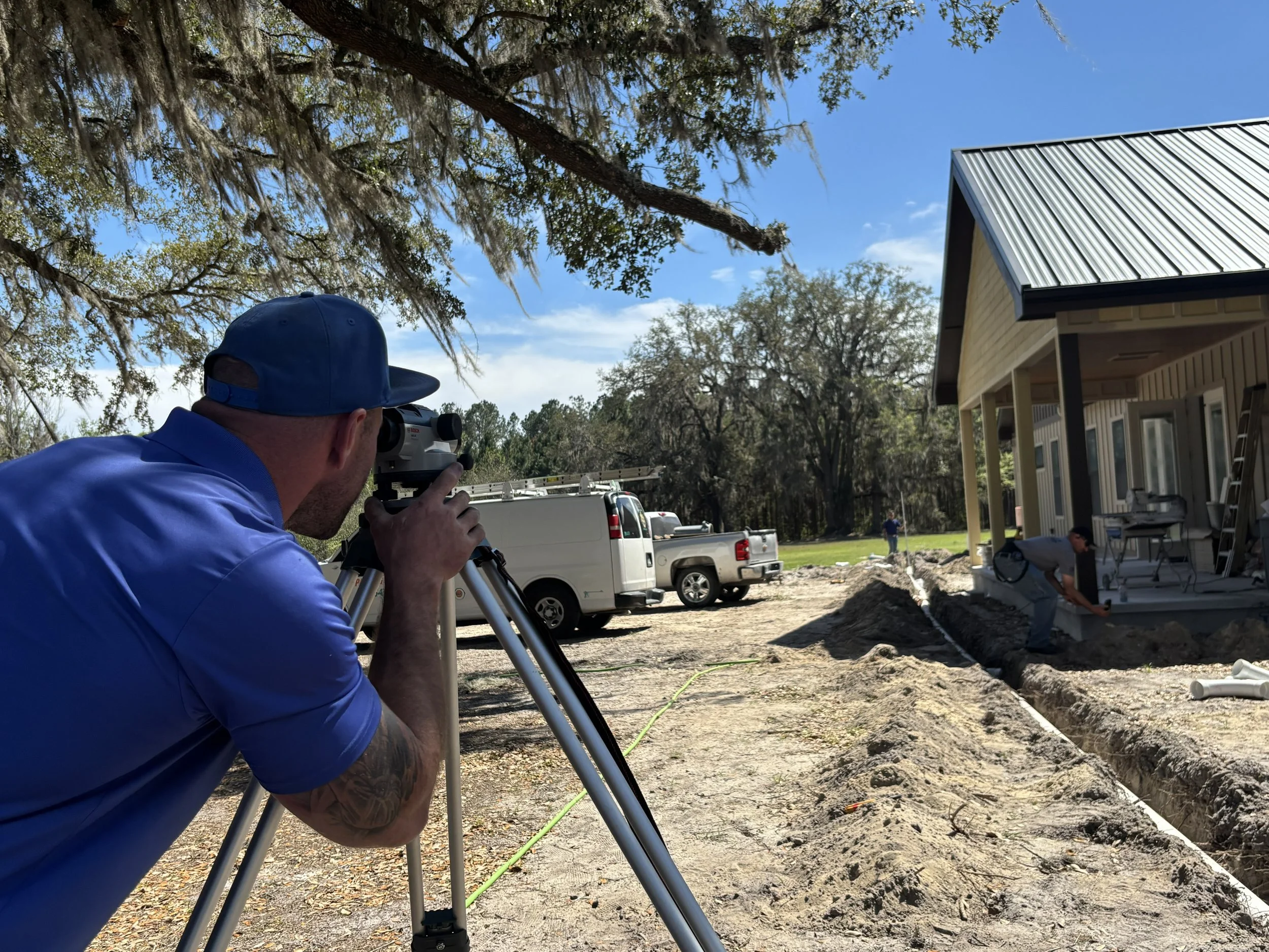 A man in a blue baseball cap and shirt using a theodolite on a tripod to survey a construction site outdoors. There is a house with an aluminum roof, trees, and vehicles in the background.