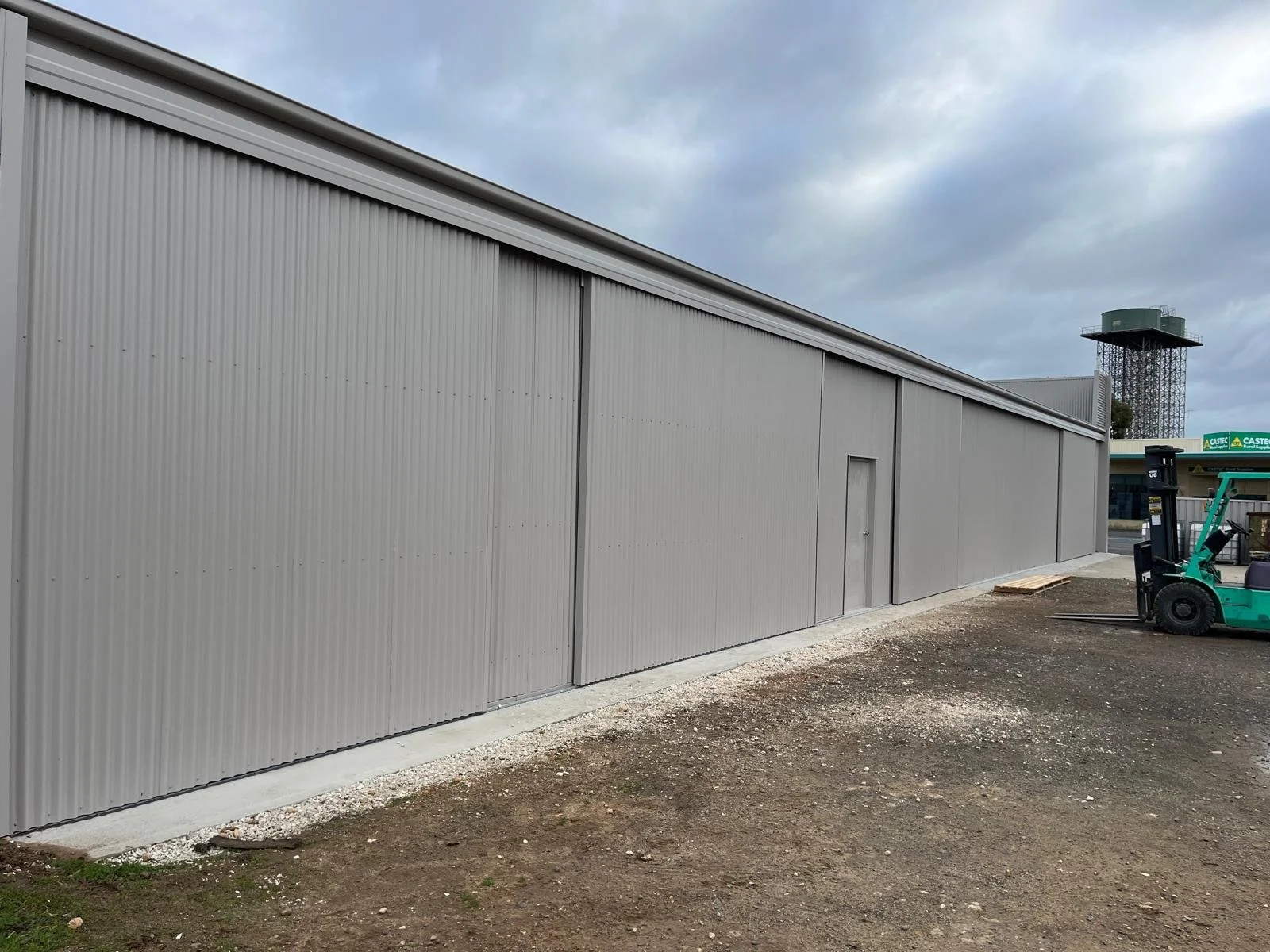 Gray metal building with vertical siding and a small door, construction site with a forklift and gravel ground, cloudy sky.