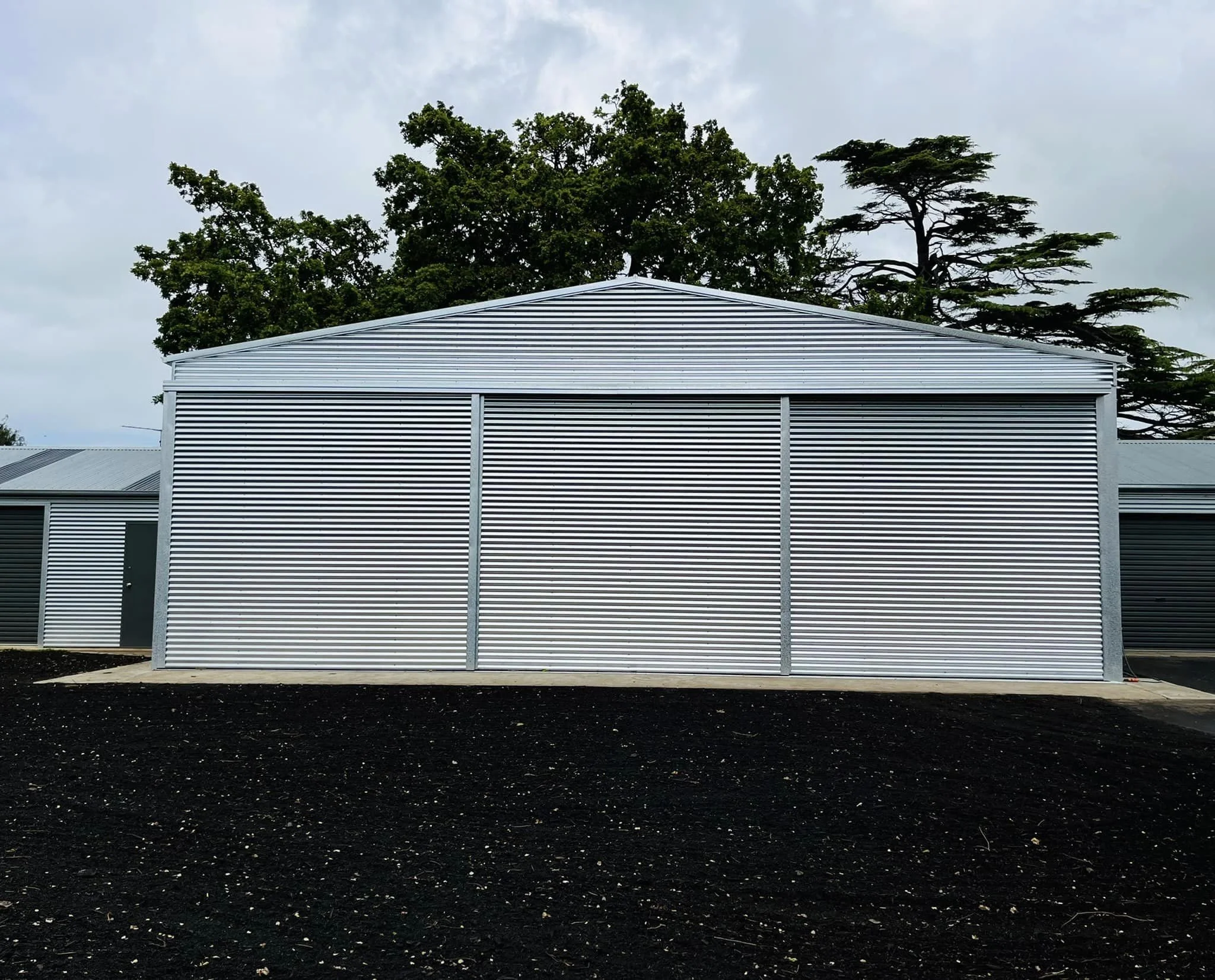 A modern building with horizontal white siding and large windows, set against a background of tall green trees and an overcast sky.