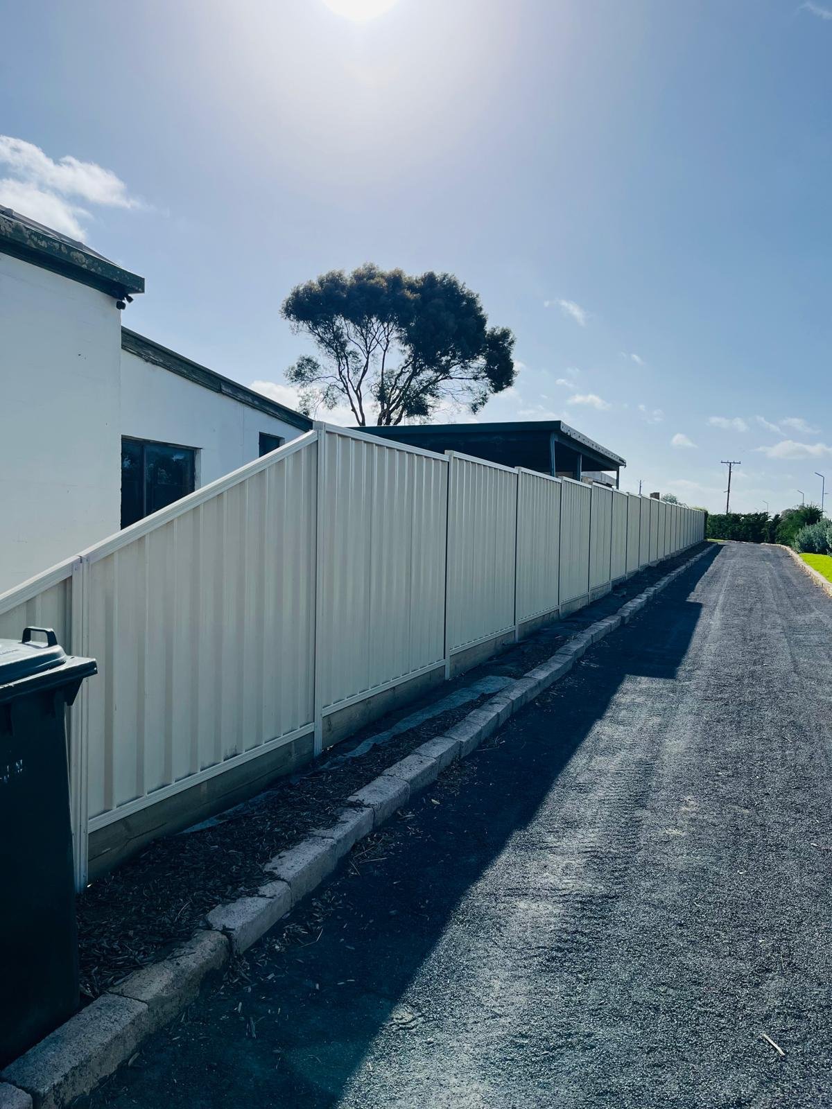 Residential street with a blacktop road, a white fence running along the side, a trash bin, a white building with windows, and a large tree in the background beneath a mostly clear sky.