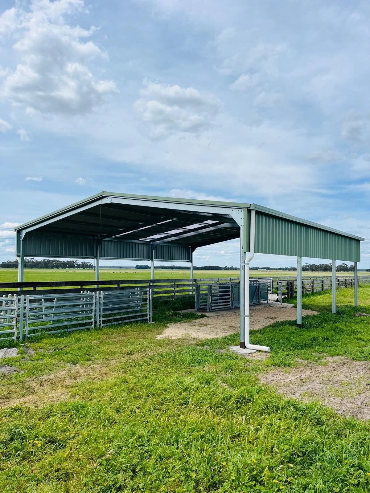 Metal structure with a green roof, used as a shelter or animal pen, situated in a grassy field with open sky in the background.
