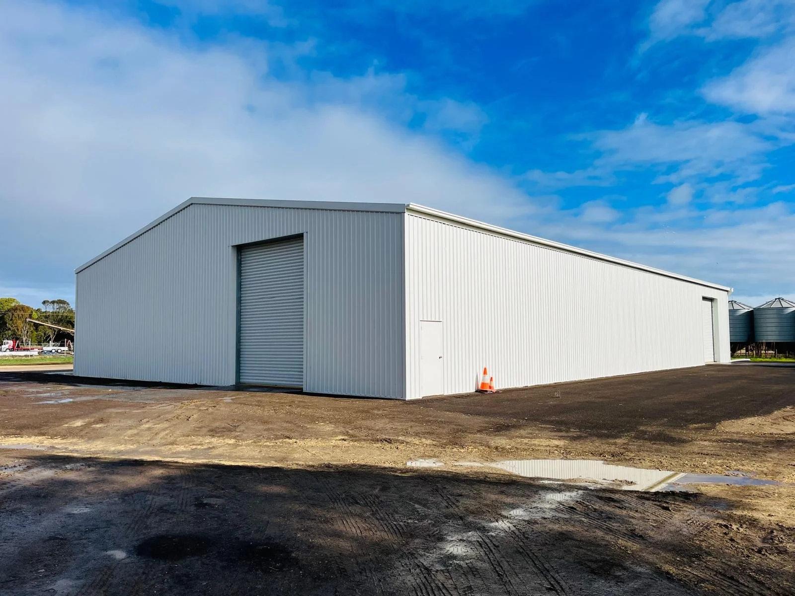 A large white metal storage building with two roll-up doors and a small side door, situated on a dirt surface with a partly cloudy sky above.